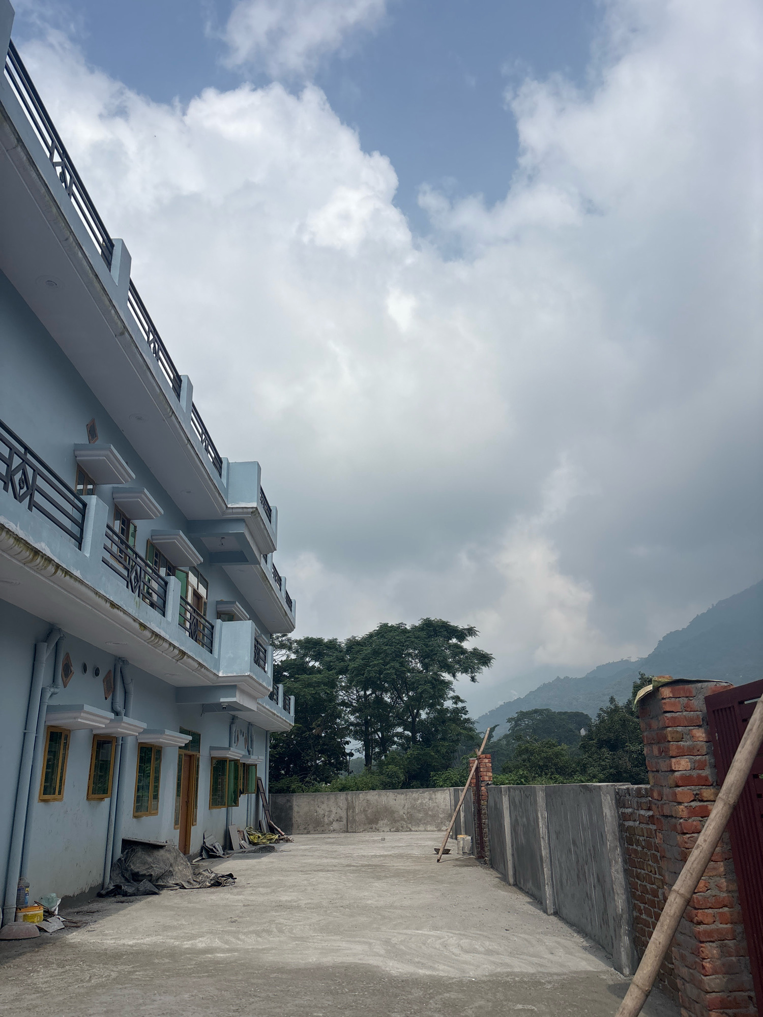 The image depicts a rooftop terrace in Bhatta, India, with a pale blue, multi-story building on the left. The building features balconies with black railings on each level, and numerous windows with yellow frames. The terrace itself is paved with concrete and appears to be under construction or renovation, with some materials visible, including a large black tarp, buckets, and construction debris.
To the right of the building, a low concrete wall runs horizontally across the frame, separating the terrace from an area with lush green trees and hilly terrain in the background. The sky is overcast with large, diffused clouds. A brick structure, possibly part of a gate or boundary wall, is visible in the far right corner, with a wooden pole leaning against it. The overall scene suggests a residential or commercial building in a semi-urban or rural setting, with a backdrop of natural landscape.