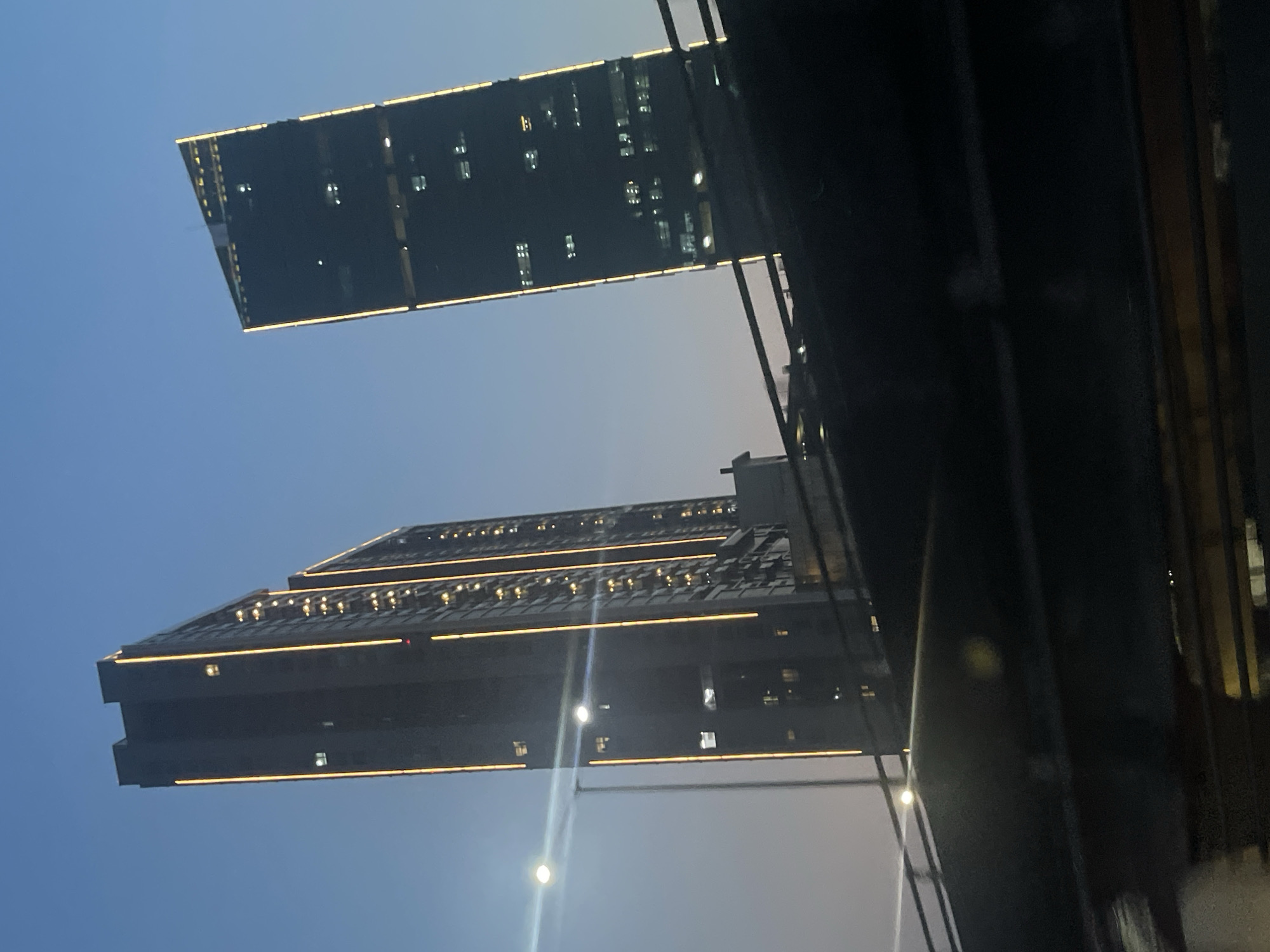 The image depicts a nighttime or dusk scene in East Jakarta, Indonesia, focusing on two prominent skyscrapers illuminated by vertical yellow lights along their edges. The sky is a clear, deep blue with a gradient of lighter hues near the horizon, indicating twilight. In the foreground, a dark, blurred structure, possibly an overpass or a bridge, partially obscures the view of the buildings. The skyscraper on the left is taller and appears to be rectangular, with rows of lit windows visible. The skyscraper on the right is slightly shorter and has a more angular, almost triangular, top, also featuring illuminated windows and edge lighting. A streetlight with a bright glare is visible to the left of the taller building. The overall atmosphere is urban and modern, with the artificial lighting of the buildings contrasting with the natural twilight sky.