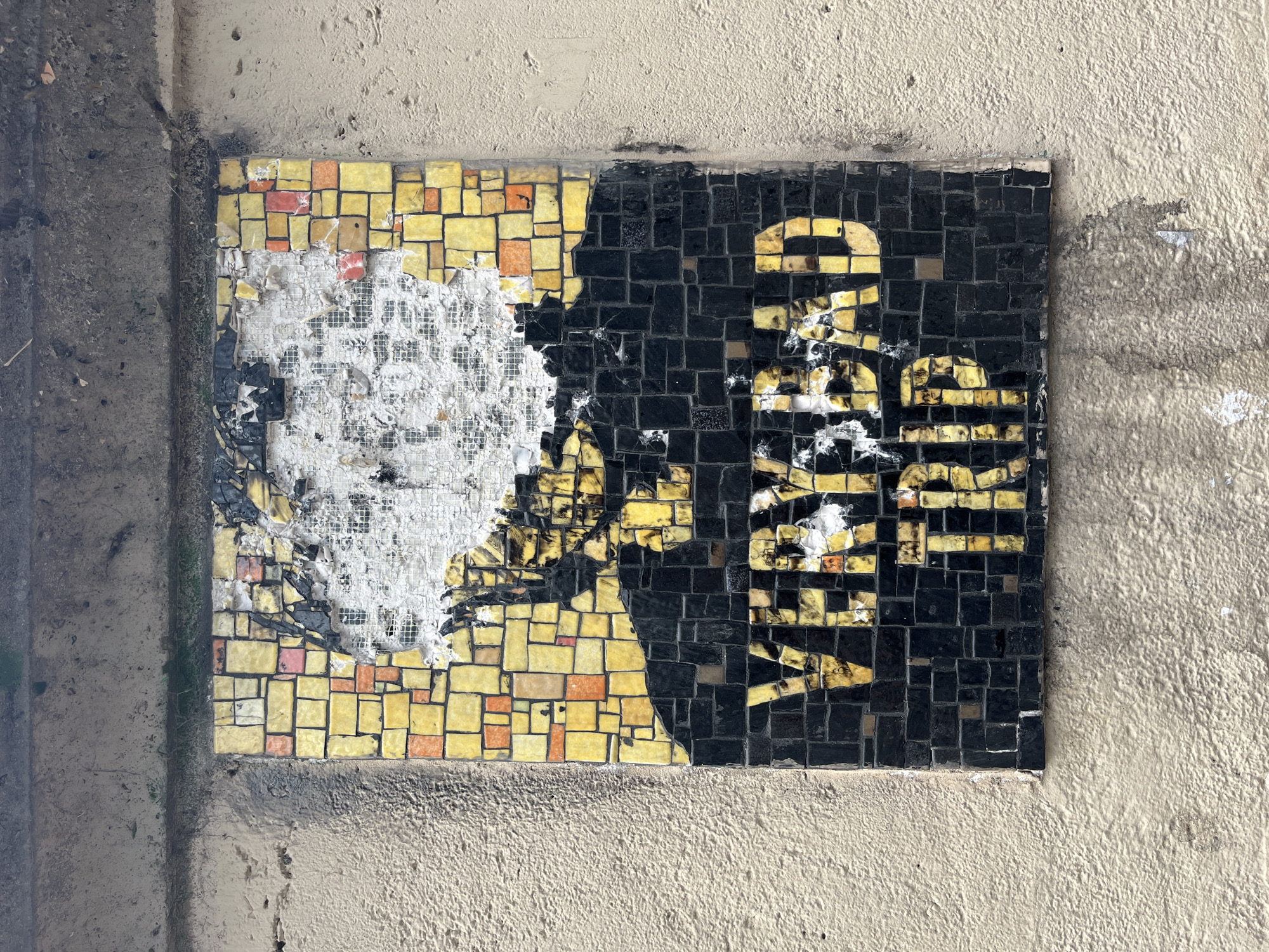 An outdoor mosaic artwork is affixed to a textured, light-colored concrete wall. The mosaic is rectangular and appears to be aged and damaged, with many tiles missing or dislodged, revealing a white mesh backing in some areas. The mosaic is composed of predominantly black tiles forming a silhouette-like shape at the bottom and yellow tiles creating text. The visible text reads "VERY BAD TRIP" in all capital letters. The yellow tiles used for the text have variations in color and some appear to be broken or chipped. Above the text, the mosaic transitions to a pattern of yellow and orange tiles, with a large section in the upper portion missing, exposing the white mesh and some darker, underlying material. The wall itself shows signs of wear and has some staining and peeling paint or material below the mosaic. The top edge of the image shows a darker, horizontal band, possibly a ledge or another architectural element.