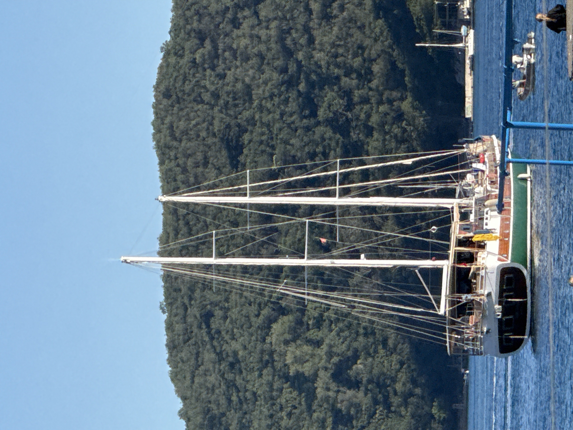 This image depicts a two-masted sailing yacht, consistent with a traditional gulet, moving across calm blue water under a clear sky in what is identified as the city of Budva, Montenegro. The large vessel, the primary subject, features a dark green lower hull, a white or light grey mid-hull, and a dark, possibly black, transom. Its two white masts are prominent, adorned with numerous rigging lines and spreaders. A small red flag is visible on the rigging of the aft mast. A yellow object, possibly a fender or bag, hangs from the starboard side near the stern. One person in a yellow garment is partially visible on the main deck, starboard side, near the base of the main mast.
In the background, a large hill covered densely with dark green trees and foliage dominates the horizon, extending from the left to the right of the frame. The sky above is a uniform, clear light blue.
In the middle ground, to the right of the main yacht, a smaller inflatable boat (RIB) with an outboard motor is visible on the water, carrying at least two individuals. Further back on the right, other smaller sailboats and a white dock or breakwater structure are present along the coastline.
In the immediate foreground, a blue metal railing cuts across the lower part of the frame on the right. To the far lower right, a person with dark hair, wearing a dark shirt, is seated or leaning on a low, light-colored concrete or stone structure, facing away from the camera and towards the water. The water exhibits gentle ripples, indicating either a slight current or the movement of vessels.