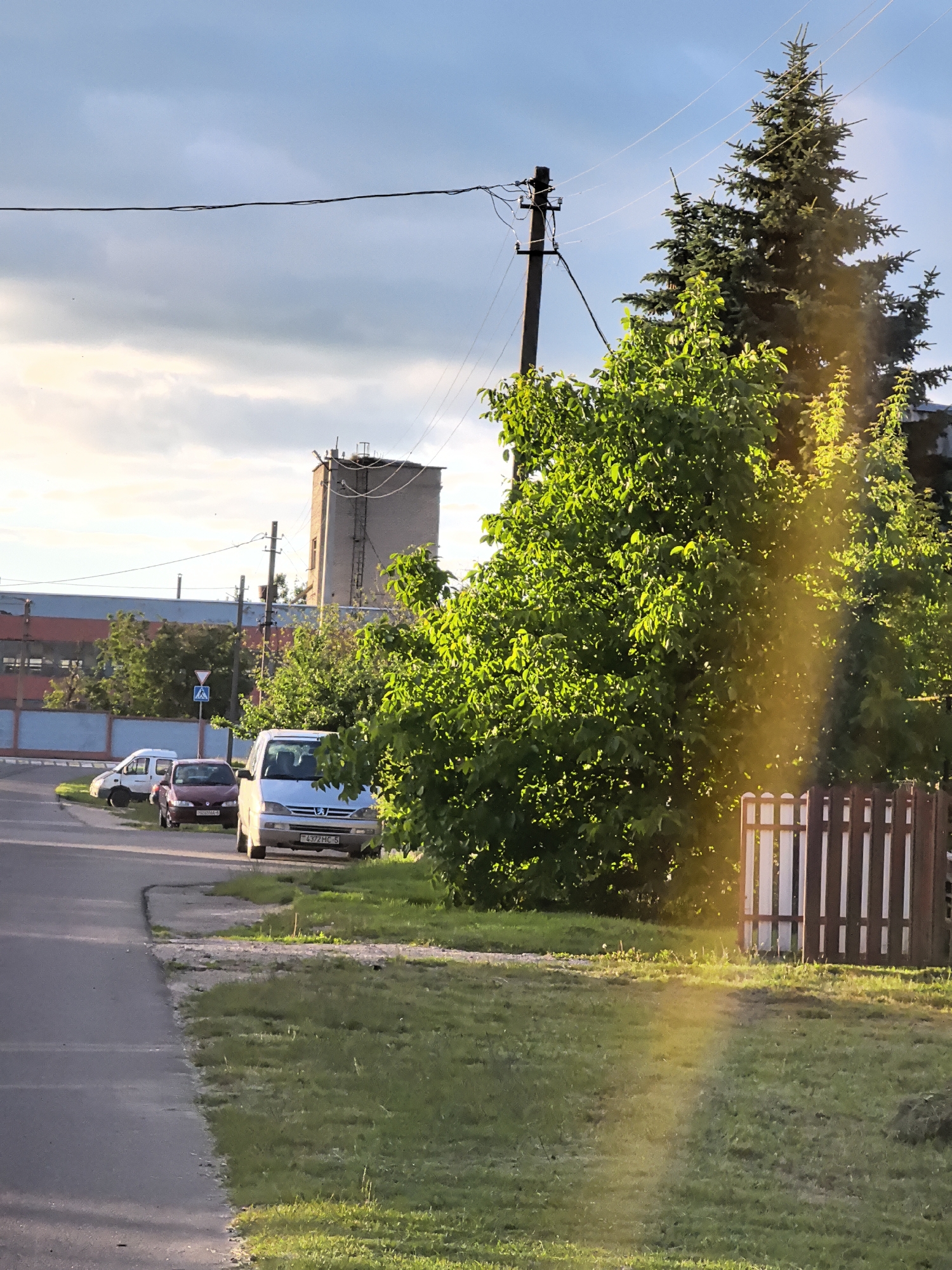 The image depicts a street scene in a suburban or industrial area. A paved road runs along the left side of the frame, bordered by a grassy verge. Several parked cars are visible on the side of the road, including a white van, a dark red car, and another white car. In the background, there is a long, low-rise building with blue and red elements, and a tall, brick industrial structure. Green trees and foliage are prominent, with a large evergreen tree on the right side of the image, partially obscured by sunlight. A wooden fence section is also visible to the right. The sky is partly cloudy, with a band of sunlight creating a lens flare effect across the right side of the image. A utility pole with wires is present in the middle ground. The overall impression is of a typical daytime scene in a developed area.