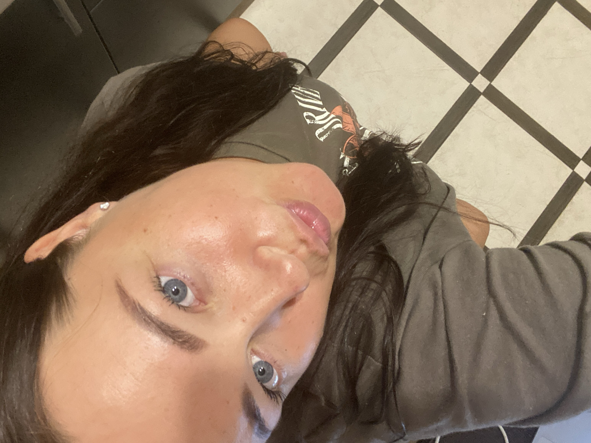 A woman with dark hair and blue eyes is shown from a high-angle perspective, looking down at the camera. She is wearing a grey t-shirt with white lettering that appears to say "DRIVE". Her lips are slightly parted, and she is wearing a small stud earring in her left ear. The background appears to be a tiled floor with a black and white checkered pattern. The scene is indoors.