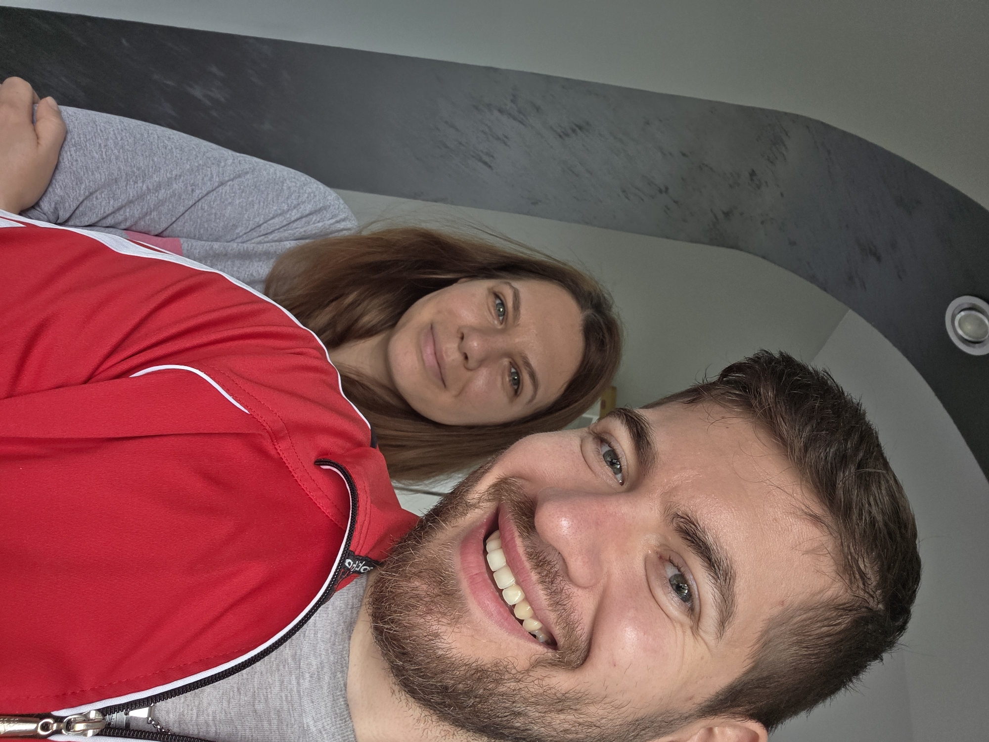 Two people are in the image. A man with dark hair and a beard is in the foreground, smiling, wearing a grey shirt and red jacket with a white stripe. Behind him is a woman with long brown hair, also smiling, wearing a grey shirt. The background is a plain wall with an arched accent stripe along the ceiling, and a recessed light fixture is visible. The setting's location in Yagodovo, Bulgaria is not discernible from the image alone.