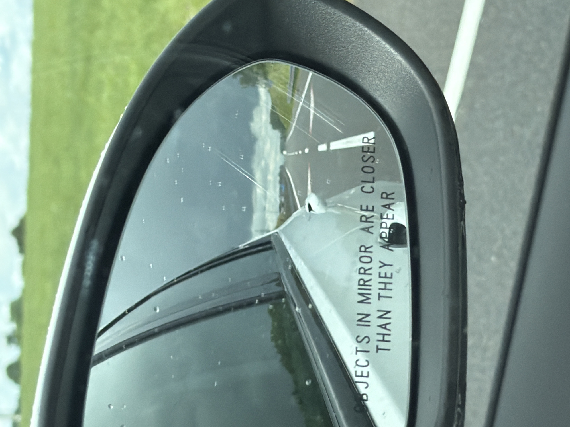 The image shows a side-view mirror of a car. The mirror reflects a highway stretching into the distance under a cloudy sky. The text "OBJECTS IN MIRROR ARE CLOSER THAN THEY APPEAR" is visible on the bottom of the mirror. The car is likely in motion on a road in a rural or suburban area, given the green fields visible in the background. There are no people or specific city landmarks identifiable in the image. The location of Пяршайскі сельскі Савет, Belarus is not evident from the visual information provided.