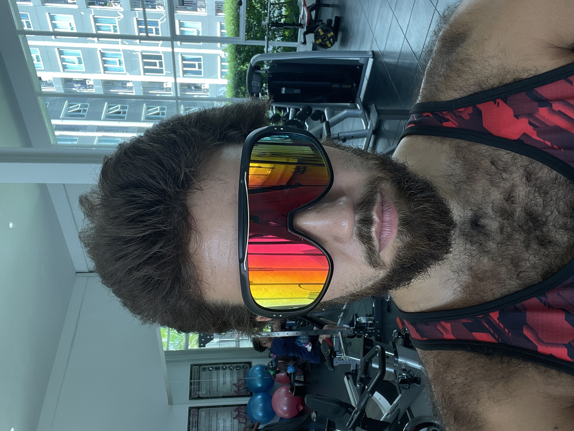 This is a close-up, front-facing, eye-level selfie of a man in a gym. He has dark brown hair, a full beard, and is wearing large, multi-colored mirrored sunglasses and a red and black patterned tank top. The background shows exercise equipment, including stationary bikes and weight machines. In the far background, through a large window, a modern apartment building with many windows is visible, suggesting an urban environment. The overall lighting is bright and appears to be natural sunlight mixed with artificial gym lighting. The man is the sole subject in the foreground, with other gym-goers blurred in the background. The scene is consistent with an indoor gym located in a city like Bangkok, Thailand, given the architectural style of the building visible.