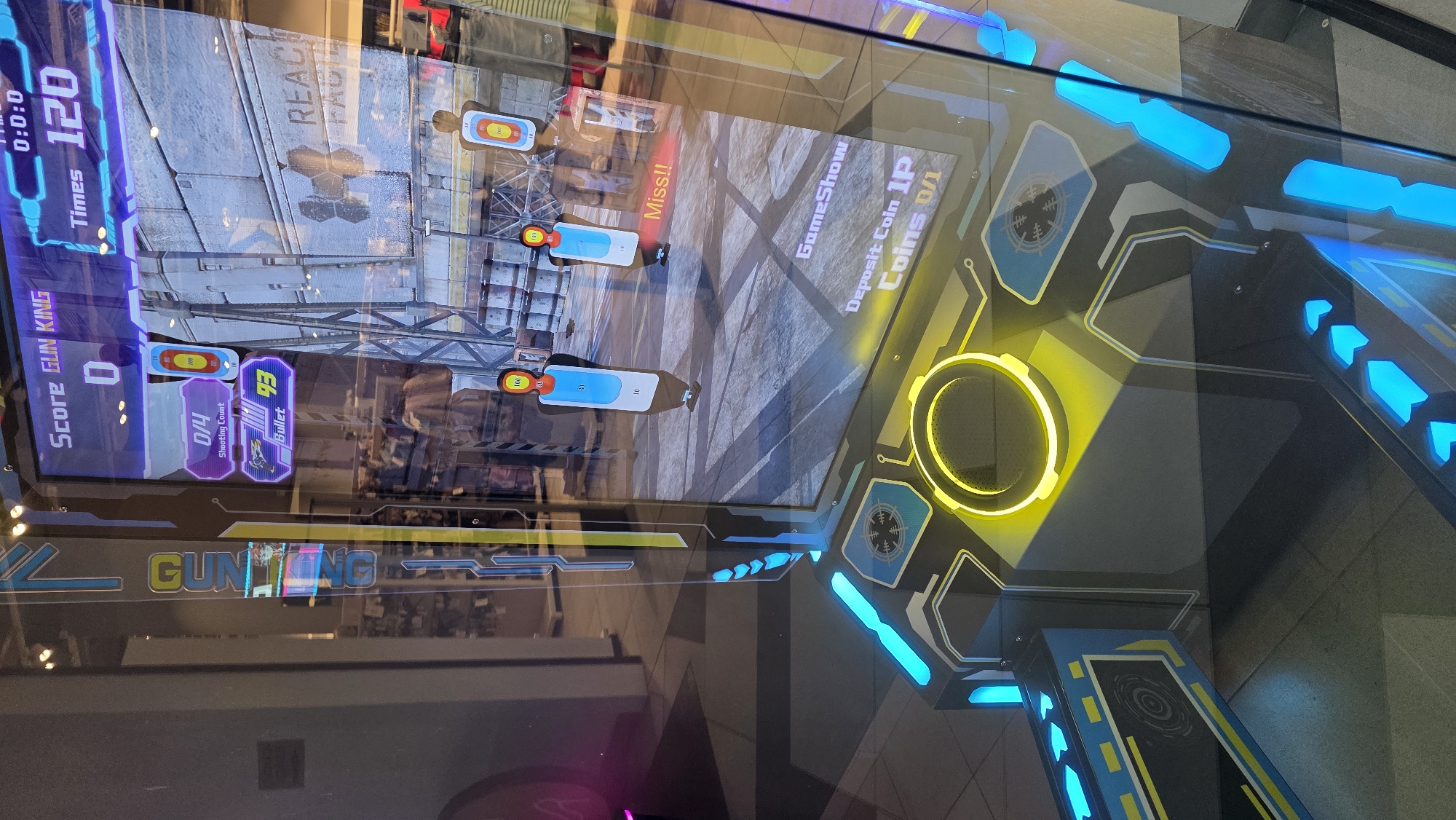 This image depicts a Gun King arcade game, likely located in an entertainment establishment, possibly in Blagoevgrad, Bulgaria, though the location cannot be definitively confirmed. The game screen displays a first-person shooter interface with a scene showing targets and ammunition counts. The game cabinet features yellow and blue neon lighting, adding to the visual appeal. Text on the screen indicates "Deposit Coin 1P, Coins 0/1," suggesting the game requires a coin to play. The word "Miss!!" is prominently displayed on the screen, indicating an unsuccessful shot.