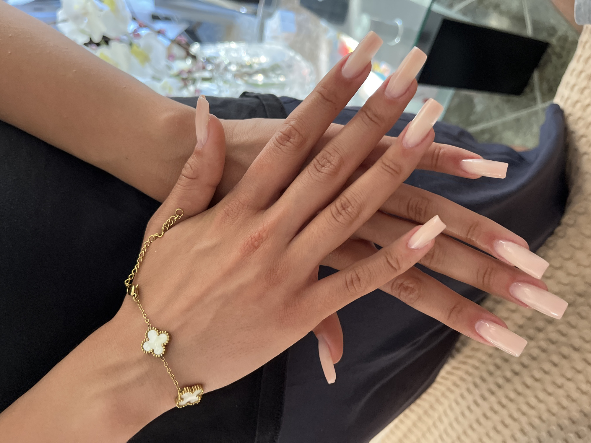 Two human hands are depicted in an indoor setting in Zlatograd, Bulgaria. The hands, displaying a light to medium brown skin tone, are positioned one over the other, with the left hand resting on top of the right. Both hands exhibit long, manicured nails with a rectangular or coffin shape, finished in a translucent light pink or nude polish. A gold-toned chain bracelet adorns the left wrist, featuring three distinct clover-shaped charms with white, possibly mother-of-pearl, inserts. The individual is wearing a dark-colored garment on the upper body and a lighter dark garment on the lower body. In the background, out of focus, a glass table is visible with reflections of white flowers arranged in a vase. A textured, light-colored surface, possibly a rug or blanket, occupies the lower left portion of the image.