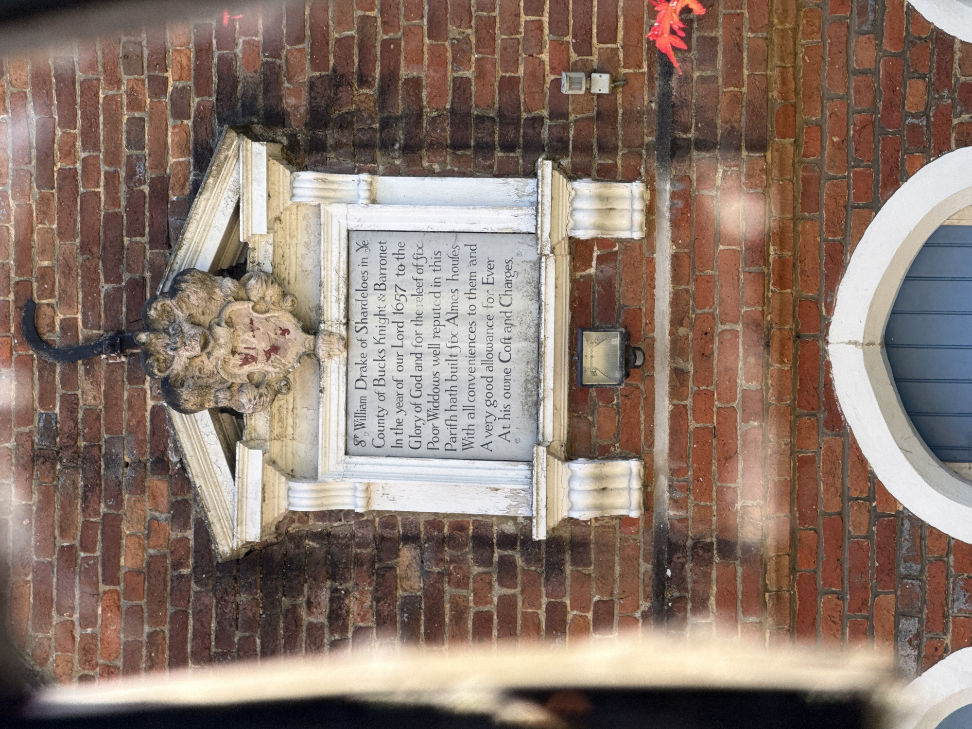 The image depicts a memorial plaque dedicated to Sir William Drake of Shardeloes, Knight & Barronet of Buckinghamshire, mounted on a brick wall, likely in Amersham, United Kingdom. The plaque commemorates Drake's construction of six alms-houses in 1657 for the relief of six poor widows of the parish, complete with conveniences and an allowance, all at his own expense. The plaque is framed with ornate carvings and is situated next to a round window. A street light is also situated on the brick wall, next to the plaque.
