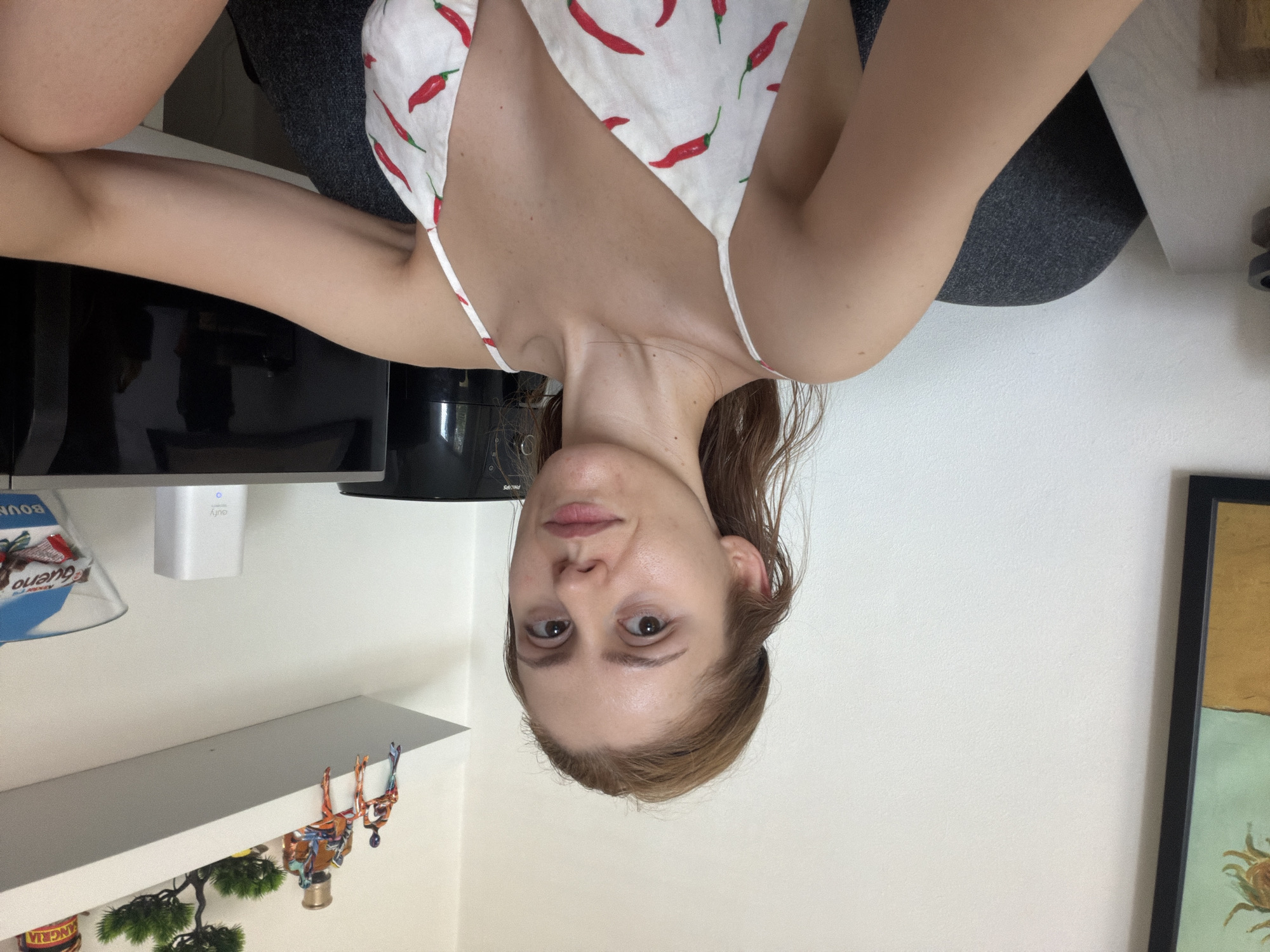The media file depicts a young woman, cropped from the chest up, with light brown hair and fair skin. She is wearing a white camisole adorned with a pattern of red chili peppers. Her head is tilted downwards, and her eyes are open and looking towards the camera. She appears to have a neutral expression. The background is a plain white wall, with hints of a dark grey surface and a shelf visible. On the shelf, there are small decorative items, including a miniature tree and some colorful figurines. A portion of a framed artwork is also visible on the far right. The lighting suggests an indoor setting, likely during the daytime. The scene appears to be a casual, personal space. There is no text clearly visible in the image.