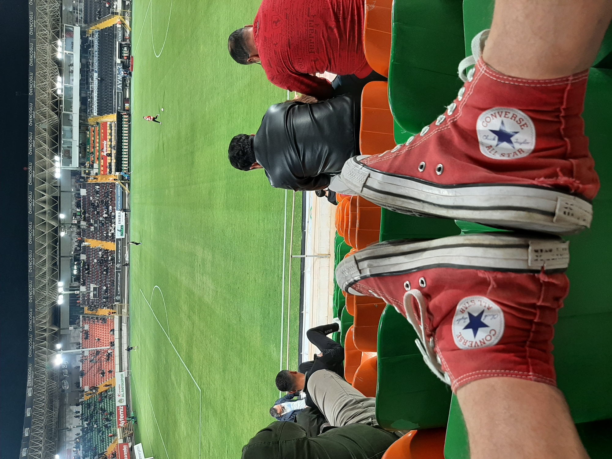 The image shows a person wearing red Converse All Star high-top sneakers, sitting in a stadium seat, presumably watching a soccer game. The stadium is filled with spectators. The soccer field is visible and green. The surrounding stadium infrastructure includes seating areas, advertising signage (Corendon Alanyaspor, Aytemiz, Anatolia Hospital, etc.) and lighting. The presence of "Alanyaspor" and the OCR-detected text "AYMER" suggests the location is Alanya, Turkey. Other people are seated nearby.