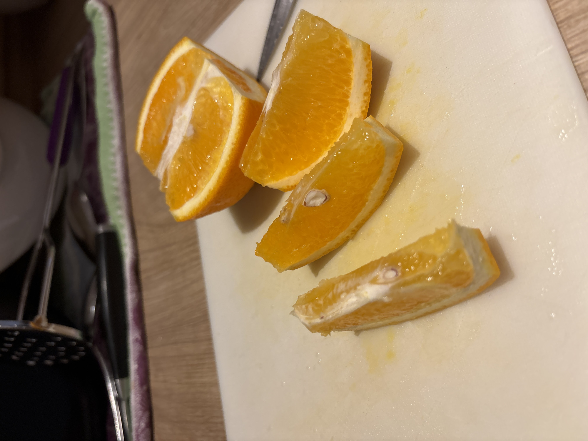 On a white cutting board are four sliced orange segments. A metal knife rests between the first and second orange segment. To the left of the cutting board, partially visible, is kitchenware. The scene takes place indoors. There is no evidence to suggest the location is in Budva, Montenegro.