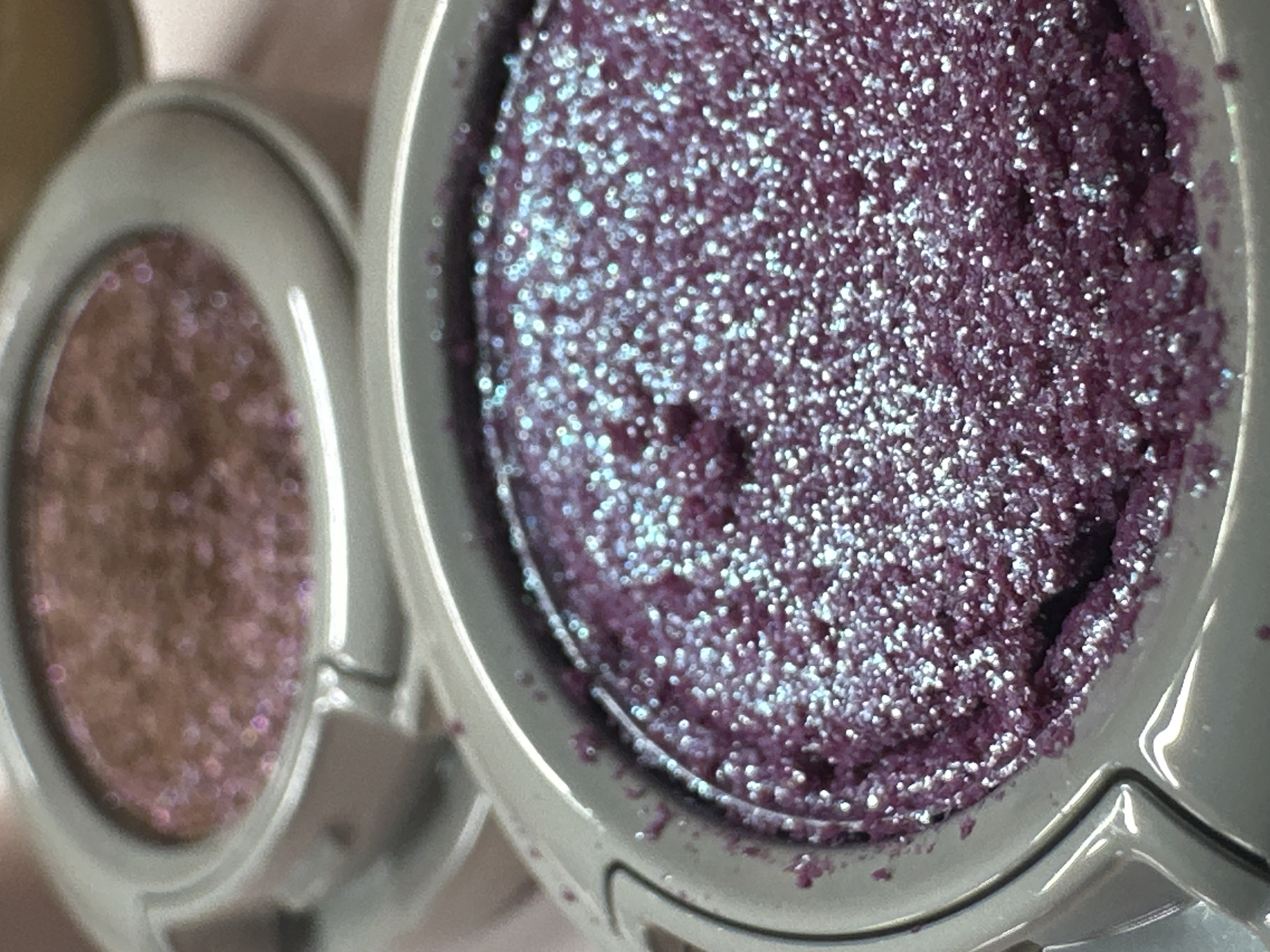 This close-up image features two open pots of glittery makeup pigment or eyeshadow. The foreground is dominated by a sharply focused circular container holding a vibrant, deep purple or magenta-colored product. This product is densely packed with fine, iridescent blue and silver glitter particles, giving it a highly shimmering and almost wet appearance. A subtle indentation or disturbance is visible in the surface of the purple pigment, suggesting it has been touched.
To the left and slightly behind, another circular pot of makeup is visible, appearing out of focus. This secondary product is a brownish-pink shade, also infused with glitter, though its texture is blurred. Both products are housed in light grey or silver-toned circular containers.
The background is softly blurred, displaying indistinct light, warm hues. The image focuses intensely on the texture and sparkle of the makeup products, highlighting their reflective qualities. No people or specific activities are depicted, nor is any text visible. While the image itself contains no explicit location cues, the scene could be situated in North Kuta, Indonesia, as per the provided context.