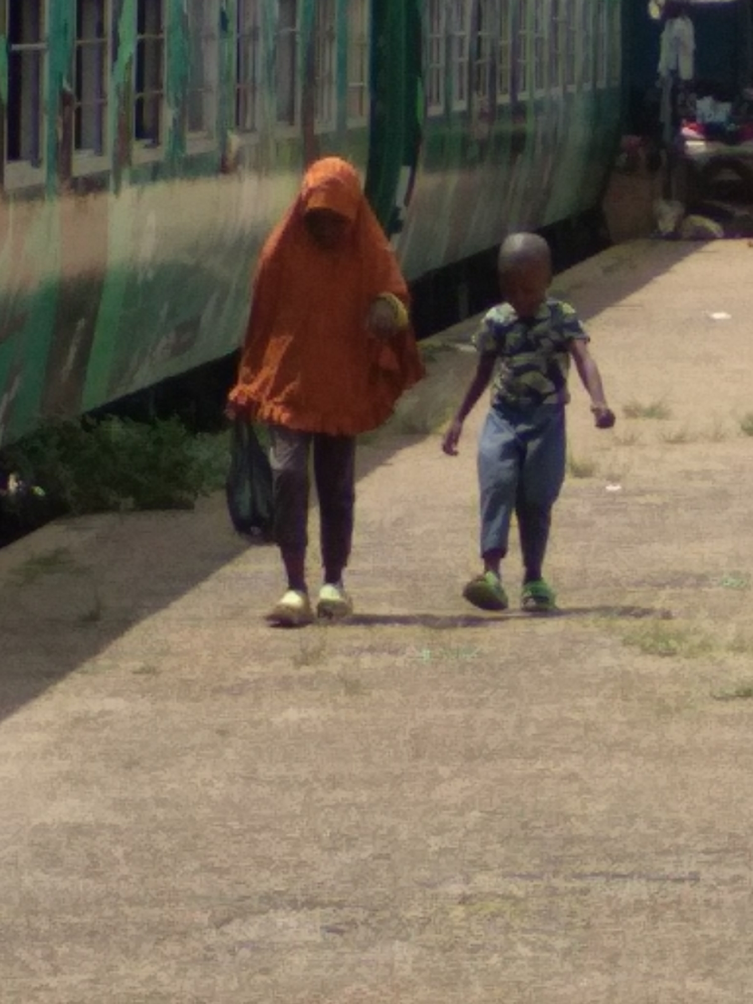 The image shows a woman and a child walking along a platform next to a train. The woman is wearing an orange headscarf and carrying a black bag. The child is wearing a patterned shirt and blue pants. The train is green and white. The location is in the city of Jos, Nigeria.