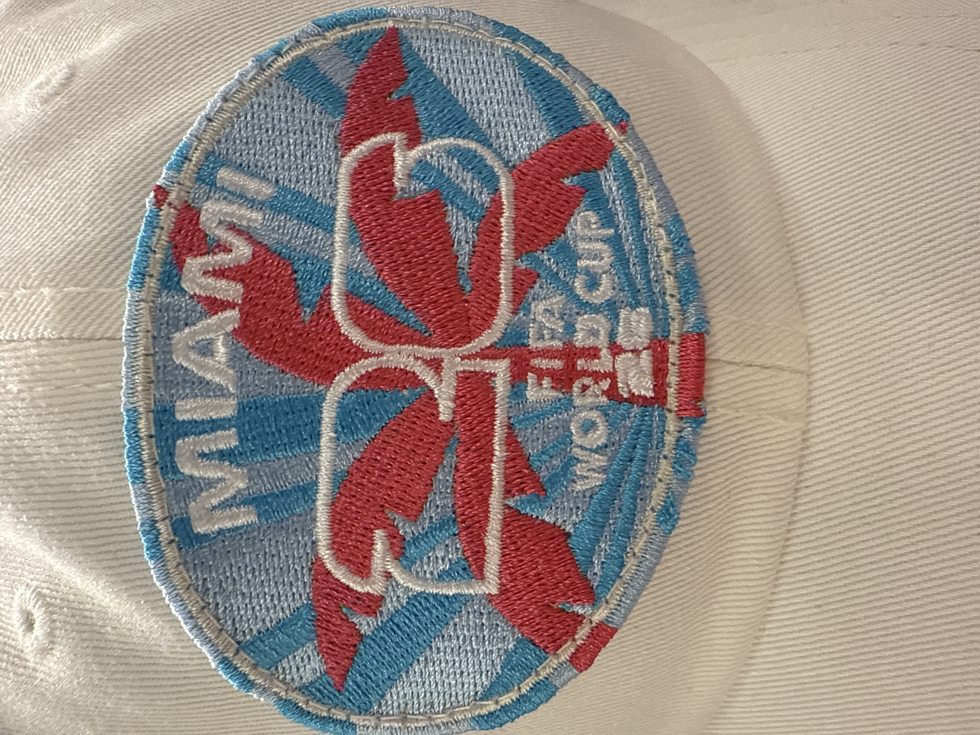 A close-up shot depicts a white baseball cap, featuring an oval-shaped embroidered patch. The patch's design incorporates the word "MIAMI" in white lettering along the left side. Centrally, stylized red palm tree graphics intersect with white outlined letters. The background consists of alternating light blue and white radiating stripes. To the right of the central design, the words "FIFA WORLD CUP" are embroidered in white. The cap's fabric appears textured, and the patch's stitching is visible. No people are visible in the image. The image is not linked to any specific location in Dania Beach, Florida.