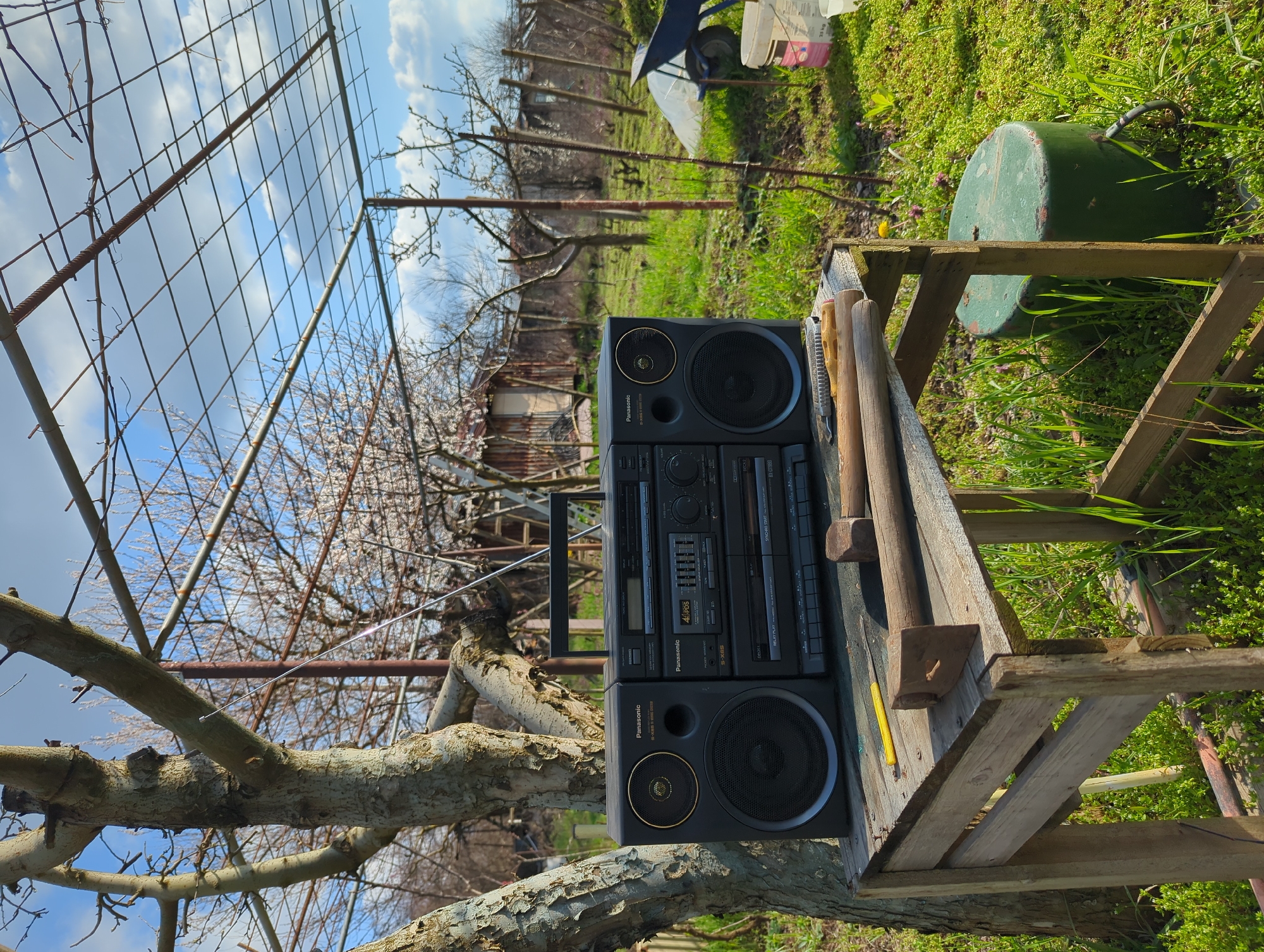 The image shows an outdoor scene in Razgrad, Bulgaria. A vintage black Panasonic boombox sits upright on a weathered wooden structure, possibly a makeshift table. Tools, including a hammer and wooden mallets, are scattered around the boombox on the table. Behind the table is overgrown grass and vegetation, along with a green cylindrical object and a garden cart. In the background, a metal fence and fruit trees with blossoms are visible. The sky is blue with scattered clouds. The boombox is centrally positioned in the frame.