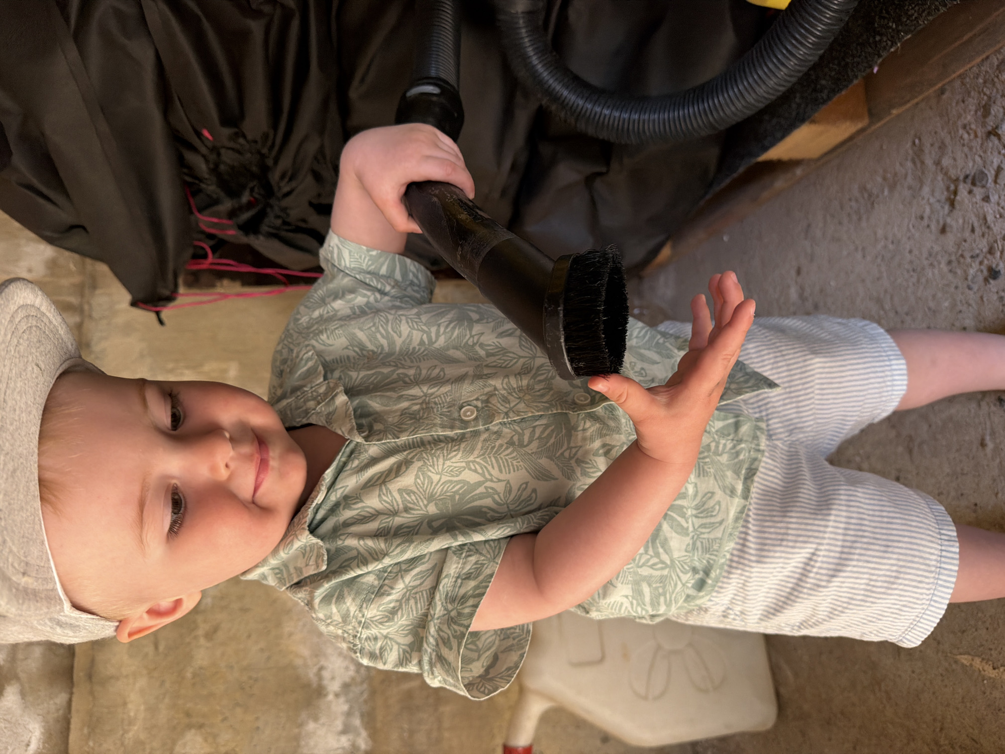 A young child, likely a baby or toddler, is lying on their back on a concrete floor. The child is wearing a light-colored hat, a light-green patterned shirt, and light-blue and white striped shorts. They are holding a black vacuum cleaner attachment in their hands. Above the child, there is a black object, possibly a bag or piece of equipment, and a black vacuum hose. The background includes a concrete wall. A white child's chair is partially visible at the bottom of the frame. The location is not identifiable as the city of Votkinsk, Russia, based on the visual information provided.