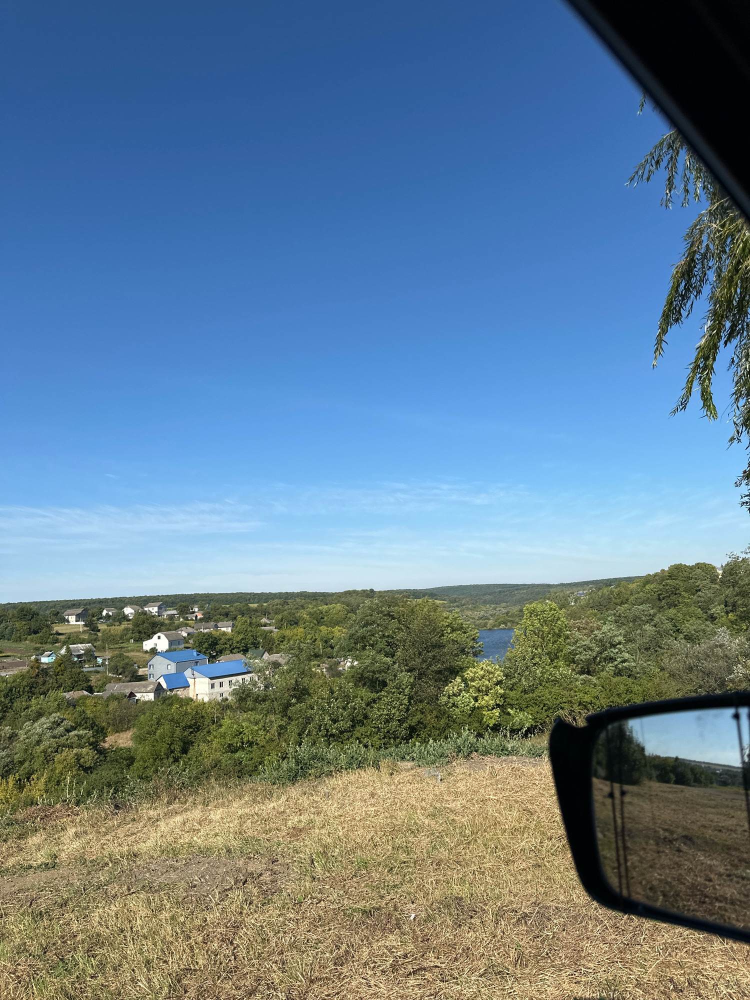 The image captures a scenic view from a car window, overlooking a village in Makiv, Ukraine. The foreground features dry, golden grass on a slope, leading down to a cluster of houses with predominantly white walls and blue roofs. A body of water, likely a river or lake, is visible in the middle ground, surrounded by lush green trees and foliage. The background displays a hazy horizon with rolling hills under a clear, bright blue sky with scattered wispy clouds. The reflection in the car's side mirror shows a portion of the landscape and trees. A branch of a tree with long, slender leaves hangs from the top right corner of the frame, adding a natural frame to the scene. The overall impression is one of a tranquil, rural setting on a sunny day.