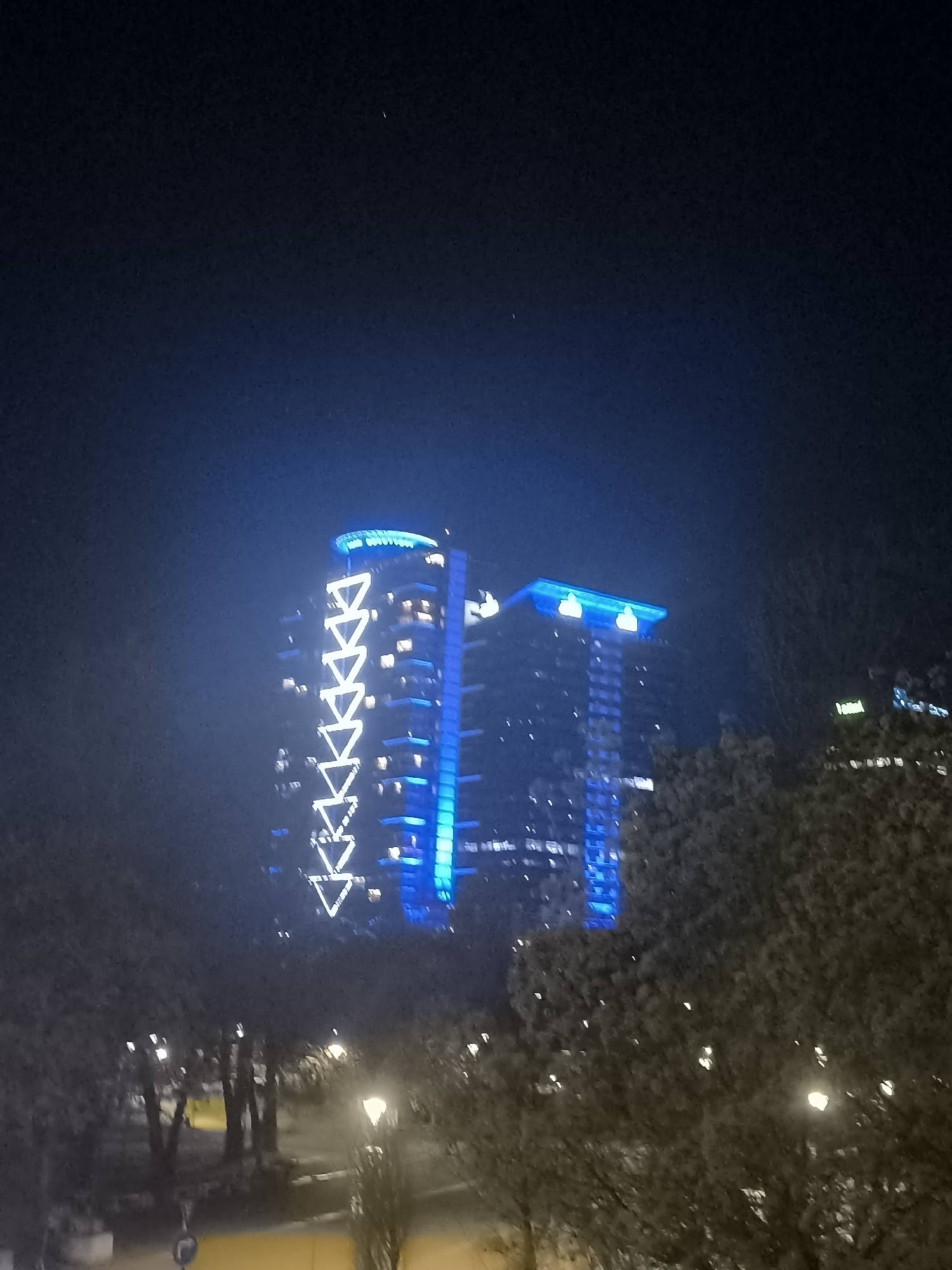 The image captures two tall buildings illuminated with blue and white lights at night, likely located in Sofia, Bulgaria. The building on the left is adorned with a vertical series of upward-pointing triangles in white light, while the building on the right has a blue-lit rectangular rooftop feature. Trees with sparse foliage are visible in the foreground, partially obscuring the view of the buildings. Streetlights illuminate a walkway or street in the middle ground, with a few indistinct figures visible in the distance. The night sky is a dark blue, creating a stark contrast with the bright building lights.