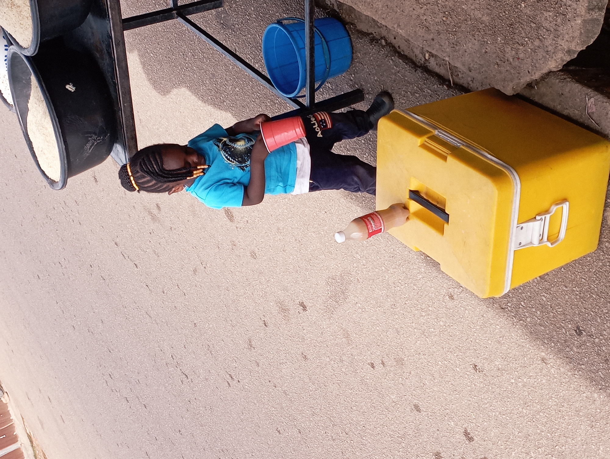 The image depicts a young child, likely a girl, standing beside a bright yellow cooler box on a paved street. The child is wearing a blue t-shirt and dark pants, with her hair styled in braids. She is holding a red plastic cup in her right hand and appears to be interacting with the cooler box. A bottle of soda rests on top of the cooler. In the background, several large black basins filled with rice are visible on a metal stand, suggesting a market or roadside stall setting. A blue bucket is also present near the stand. The street surface is asphalt, with some visible cracks and debris. The lighting suggests it is daytime, with shadows indicating the sun's position. The overall scene is consistent with a street vendor environment in Jos, Nigeria, where such displays of goods and activities are common.