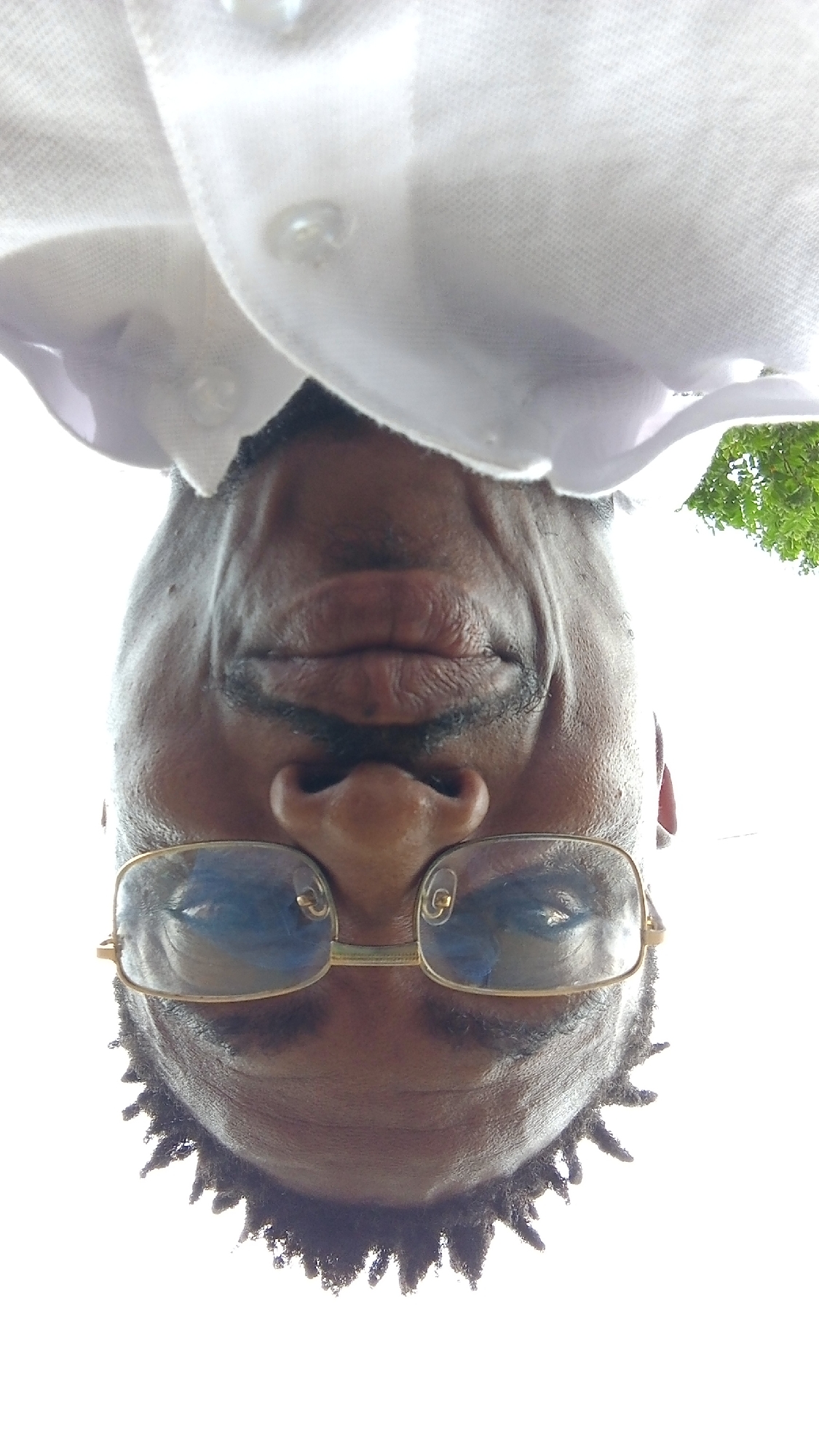 The image is a close-up, upside-down selfie of a man wearing glasses and a white collared shirt. The man's face is centered in the frame, with his chin and part of his head visible at the bottom. He has dark skin, a mustache, and short, spiky hair. His glasses have gold-colored rims and blue-tinted lenses that reflect the sky. The background is predominantly white, with a hint of green foliage visible in the upper right corner, suggesting an outdoor setting. The image appears to have been taken in Zango-ndaji, Nigeria.