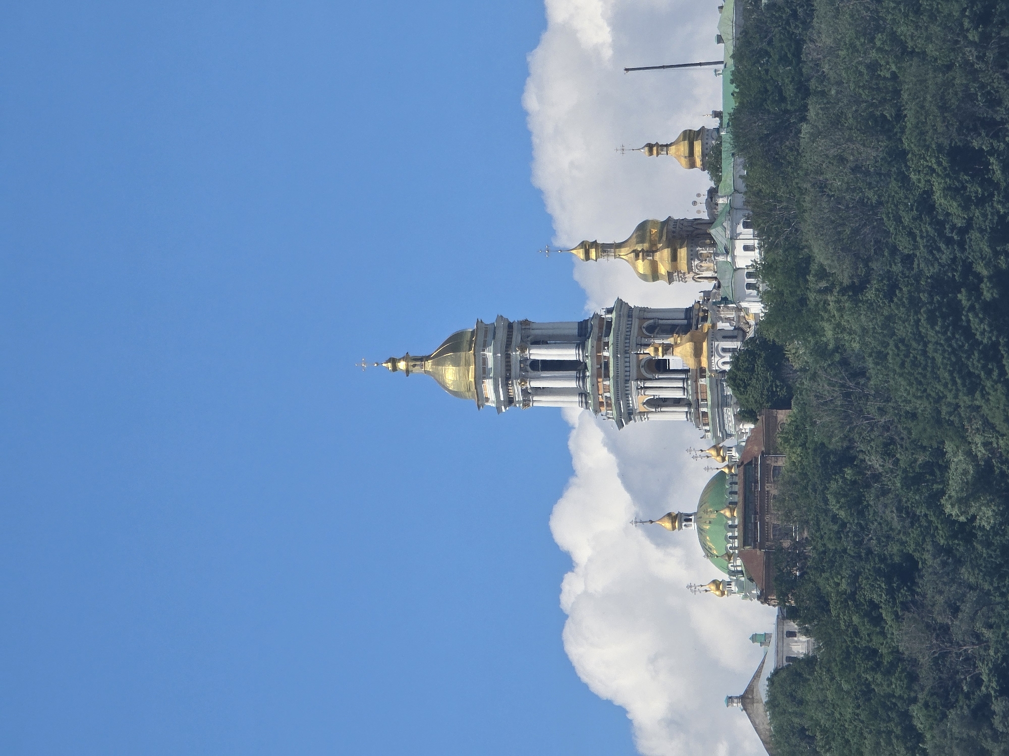 The image shows a view of the Kyiv Pechersk Lavra, a historic Eastern Orthodox monastery complex located in Kyiv, Ukraine. The monastery is situated on a lush green hillside overlooking the city. The foreground is dominated by dense trees, with the monastery's distinctive golden domes and bell tower rising above the foliage against a clear blue sky with fluffy white clouds. The architecture features multiple domes, some gilded and some green, and an imposing bell tower with intricate detailing. The overall scene conveys a sense of historical and spiritual significance, bathed in daylight under a bright sky. There are no people visible in the image.