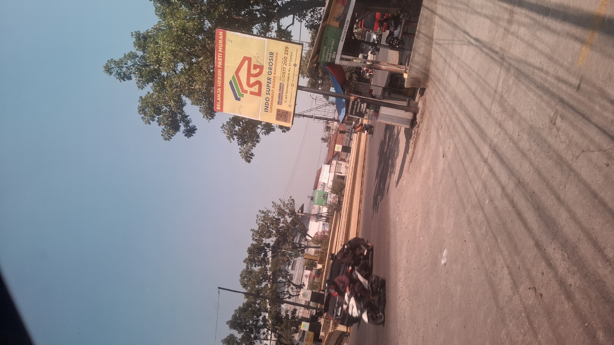 The image captures a street scene in Rancagoong, Indonesia, under a clear blue sky. A large yellow billboard for "INDO SUPER GROSIR" stands prominently on the right, advertising "BELANJA HEBOH PASTI MURAH" (Big Shopping, Definitely Cheap). The billboard also lists a phone number, 0817 200 239, and an address, Jl. Arif Rahman Hakim No. 27 Cianjur. Below it, a smaller sign for "Warung Pak Ade" is visible.
The street is paved, and several vehicles are present. Two motorcycles are in motion in the foreground, one white and one dark. A dark car is also visible. In the background, buildings and trees line the street, with some displaying the "GroBike" logo, indicating a ride-sharing service. The overall environment suggests a busy commercial area. The sunlight casts long shadows across the road, indicating it is likely afternoon. There is some scattered litter on the road surface.