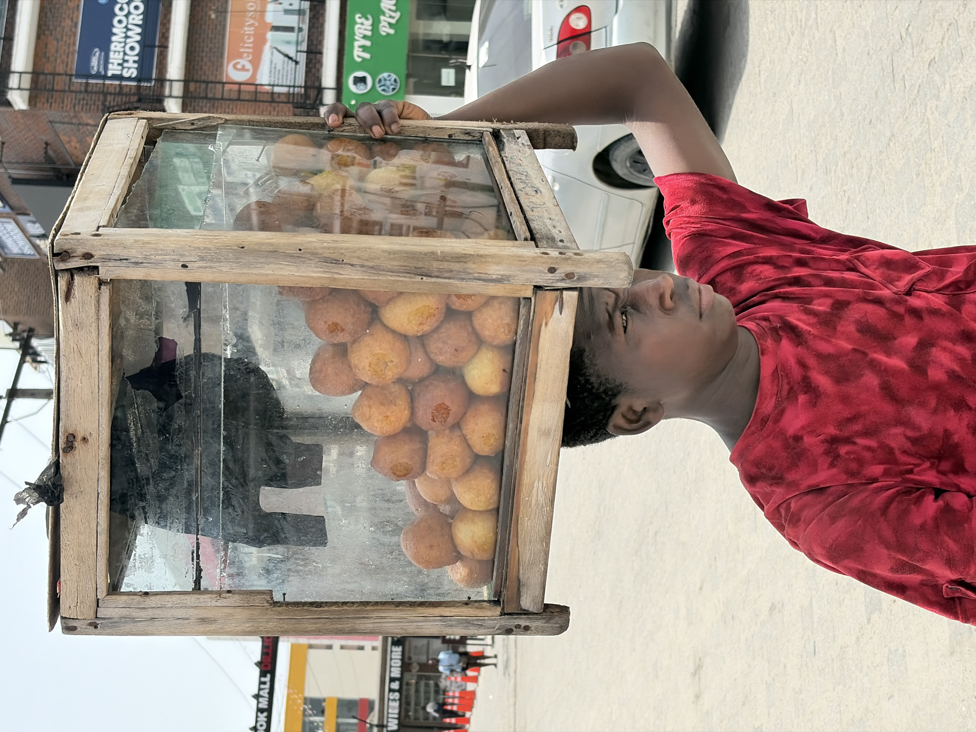 The image depicts a young man in Alaguntan, Nigeria, wearing a red t-shirt. He is standing on a paved surface, likely a sidewalk, and appears to be selling puff-puff (a type of fried dough snack) from a glass-fronted wooden display case. The case contains two types of puff-puff, one golden and the other brown. In the background are street signs and buildings. A white car is partially visible behind the man.