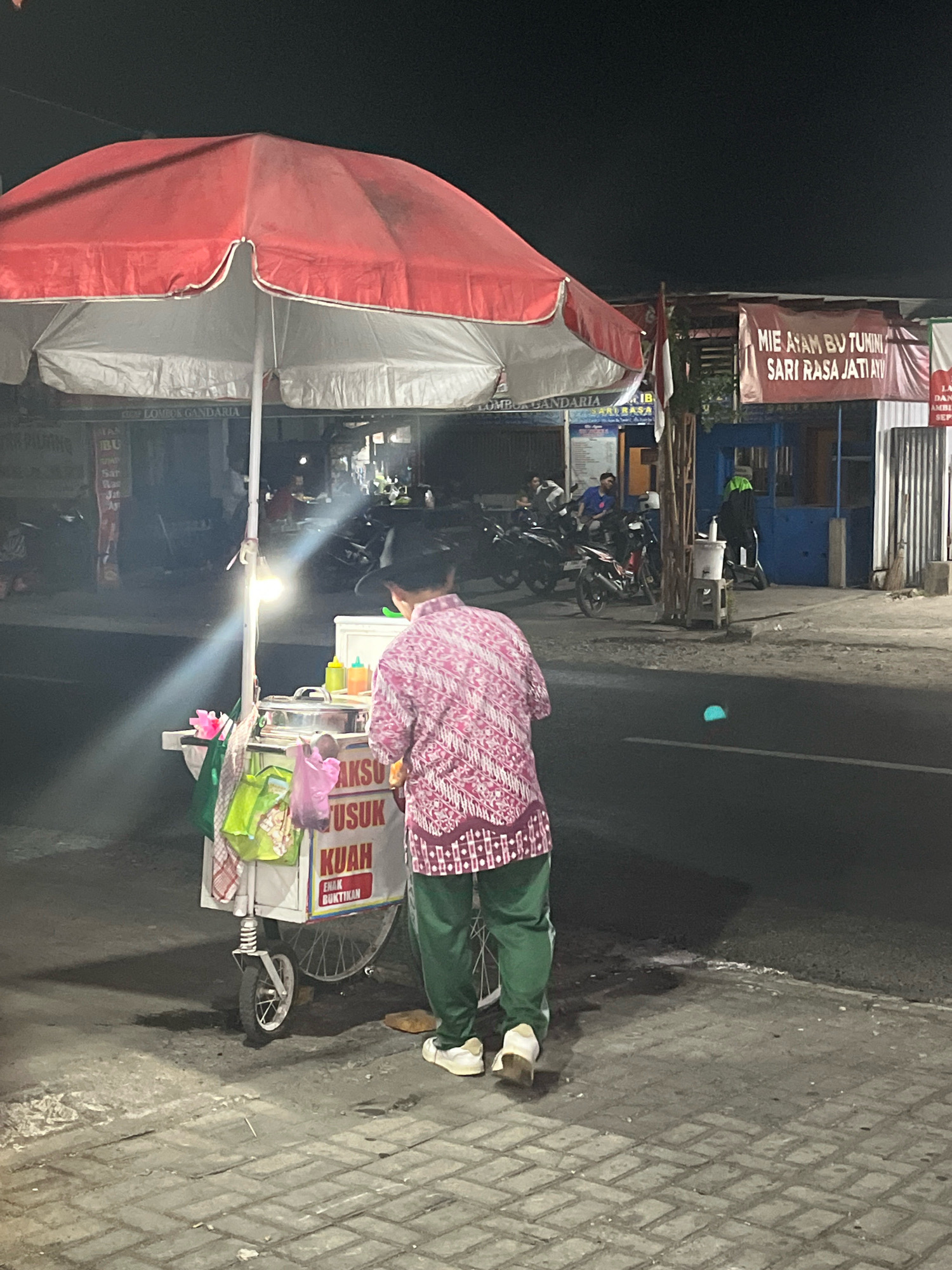A person in a pink patterned shirt and green pants stands at a food cart under a red and white umbrella at night. The cart is illuminated by a hanging lightbulb and has a sign that reads "Bakso Usus Kuah Enak Buktikan". Several plastic bags, one pink and two green, are attached to the cart. In the background, a banner reads "Mie Ayam Bu Tumini Sari Rasa Jatiayu". Motorcycles are parked in front of shops in the dimly lit street. The scene appears to be in Banguntapan, Indonesia.