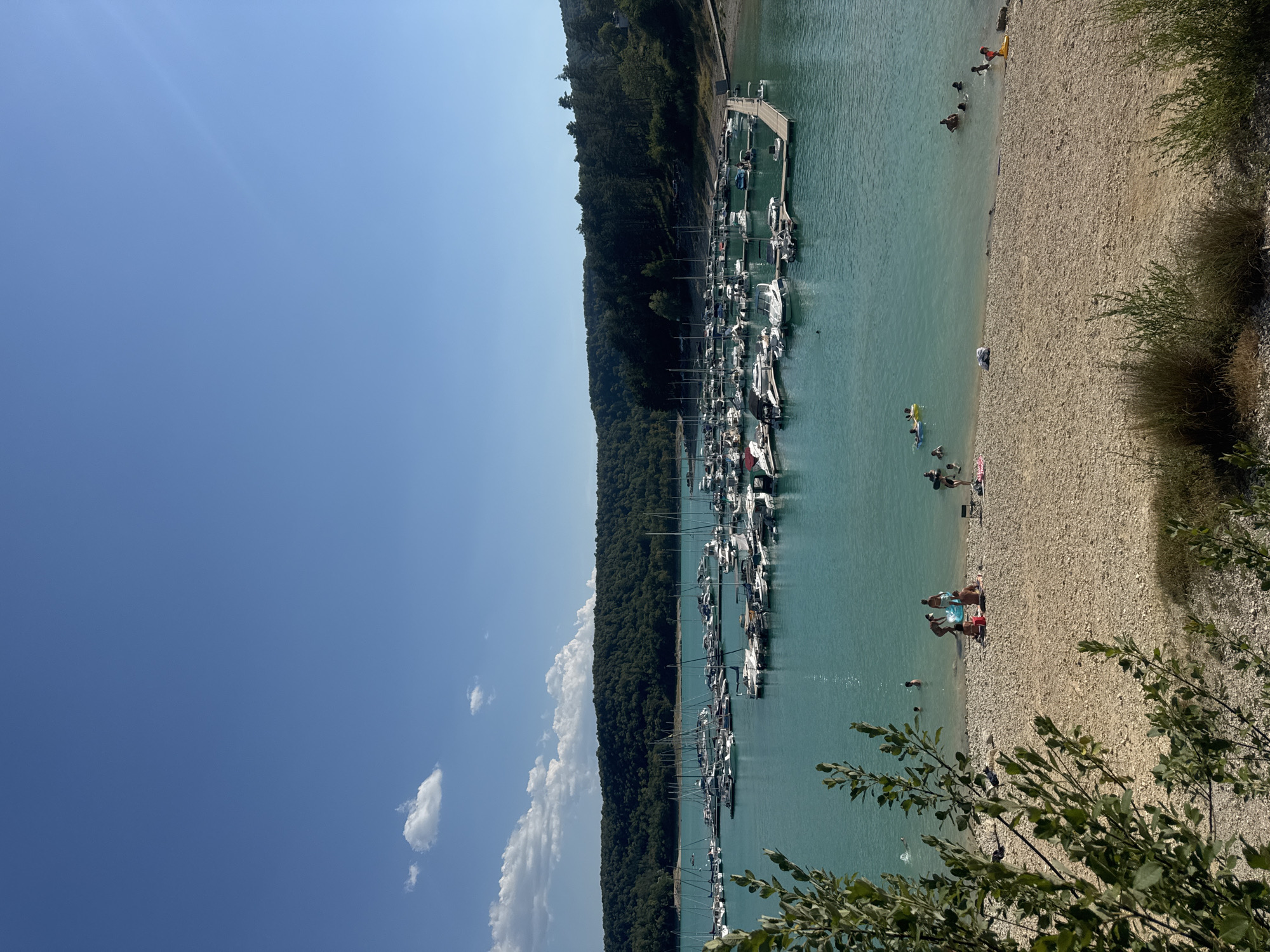 The image depicts a scenic view of a lake with a marina in Maisod, France. A vast expanse of clear, turquoise water dominates the foreground and midground, dotted with numerous boats docked in the marina. The boats, mostly white with visible masts and hulls, are neatly arranged, suggesting a well-maintained facility. A wooden pier extends into the water, with a few people visible on it and near it.
The shoreline is a mix of sand and pebbles, with some sparse vegetation growing along the edge. Several people are seen enjoying the water and the beach. Some are swimming, while others are relaxing on the shore or playing in the shallows. A child can be seen on a yellow float. The beach area has a few towels and beach chairs scattered around, indicating a leisurely atmosphere.
In the background, a lush, tree-covered hillside slopes down towards the lake, forming a picturesque backdrop. The sky above is a clear, vibrant blue with a few scattered white clouds. The overall impression is one of a tranquil summer day at a popular lakeside recreational area. The lighting suggests it is daytime, with the sun casting clear shadows.