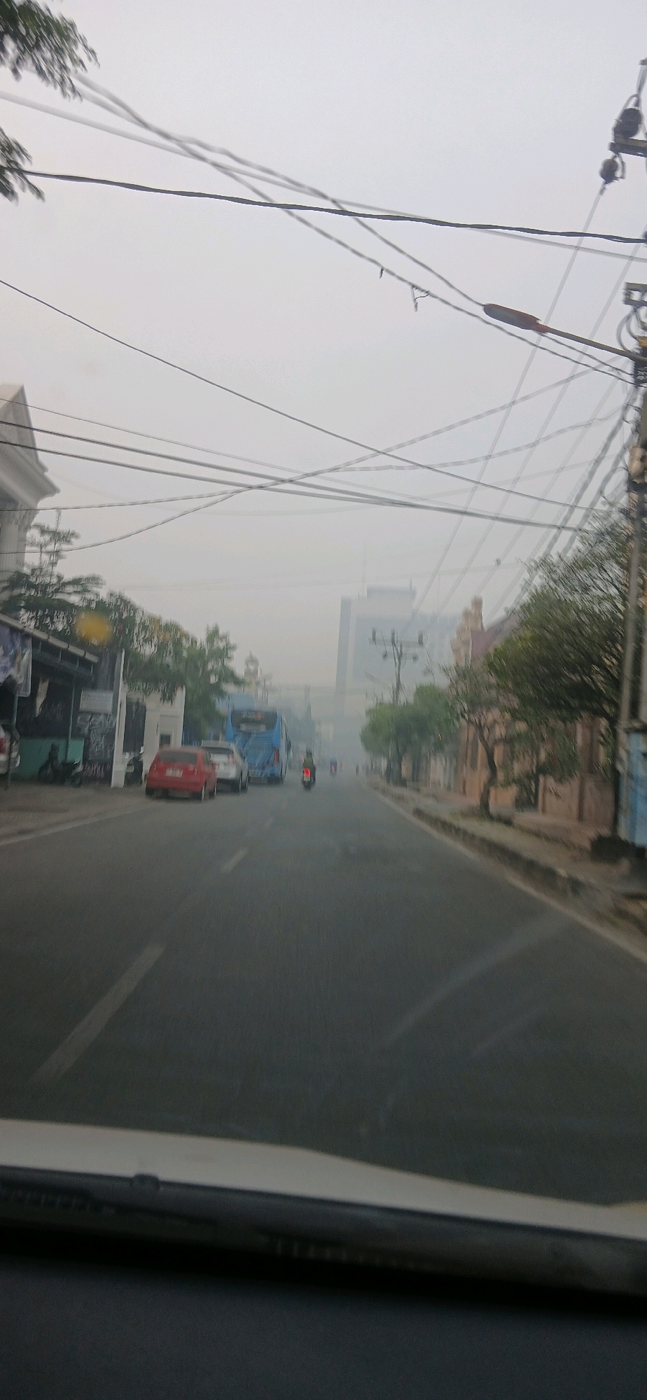 The image depicts a street scene in Samarinda, Indonesia, under a hazy or foggy sky, likely during the day. A road with lane markings is visible, with vehicles moving along it. In the foreground, the dashboard of a vehicle is partially visible, suggesting the photo was taken from inside a car.
On the left side of the road, there are buildings and trees. One building has a distinct white facade with architectural details. Further down the road on the left, a blue bus is prominent, indicating public transportation is active. A red car and a white car are parked or stopped near the bus.
On the right side of the road, there are more trees and buildings. A utility pole with numerous wires is a significant feature in the sky, indicating extensive electrical and communication infrastructure. The overall atmosphere is subdued, possibly due to the atmospheric conditions or time of day.
Several motorcycles are also visible on the road, suggesting their common use as a mode of transport in the area. The buildings appear to be of various architectural styles, some modern and others older. The presence of street signs and sidewalks suggests an urban environment. The haze obscures distant details, creating a somewhat diffused view of the cityscape.