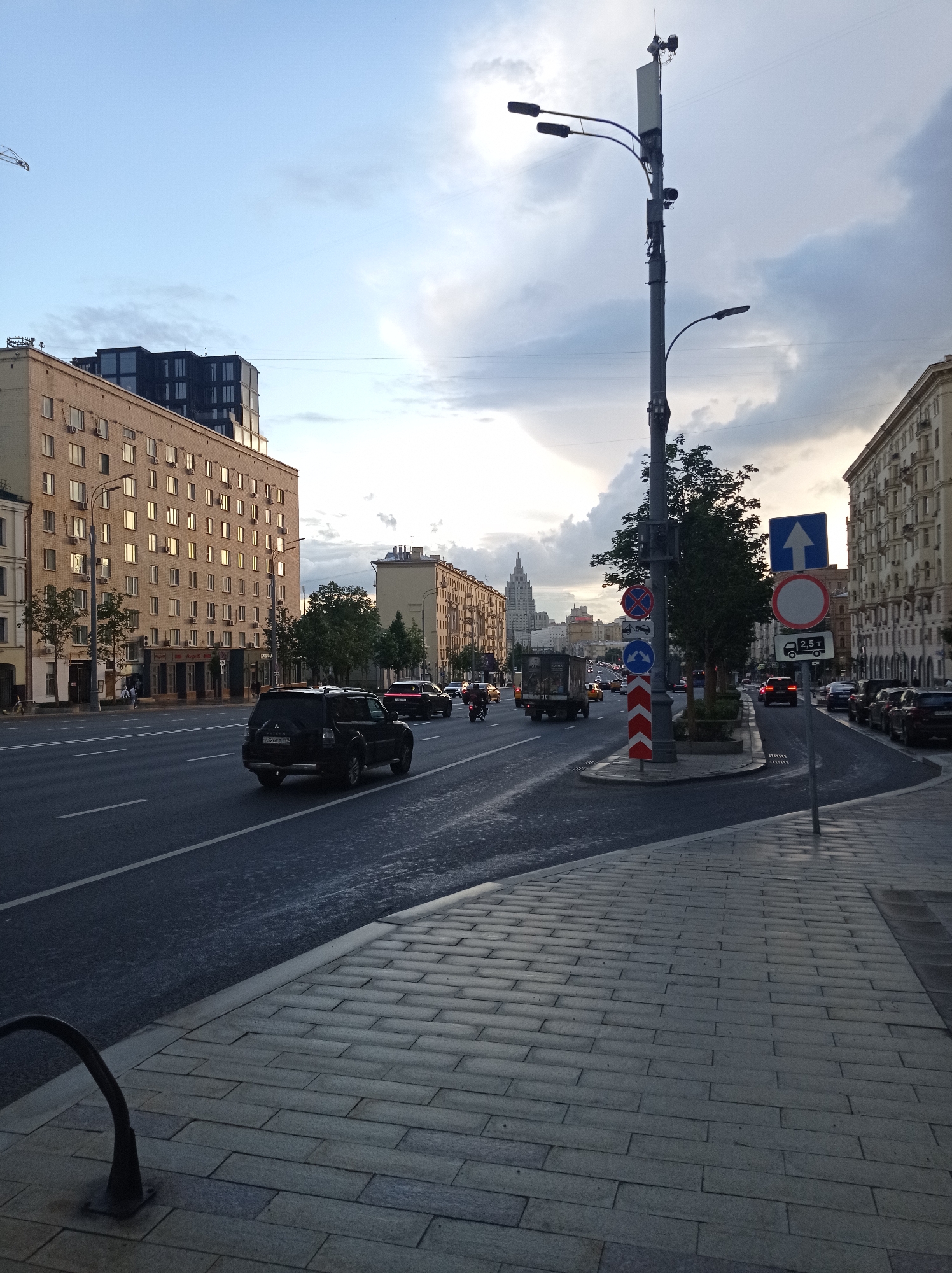 The image depicts a street scene in Moscow, Russia, likely during the daytime, with overcast skies and dramatic clouds. The perspective is from a sidewalk, looking towards a multi-lane road.
On the left side of the road, a large, beige apartment building with many windows dominates the foreground. Adjacent to it, a darker, modern building rises higher. The road itself has visible lane markings and is occupied by several vehicles, including a black SUV in the foreground, a motorcycle, a truck, and other cars. Traffic lights and road signs are present, including a sign indicating a weight limit of 2.5 tons.
On the right side of the road, another apartment building stretches into the distance. A tall lamppost with multiple lights and communication equipment stands prominently near the sidewalk. The sidewalk is paved with rectangular stones. The environment suggests an urban setting with a mix of older and newer architecture. The overall impression is of a typical city street with active traffic under a cloudy sky.