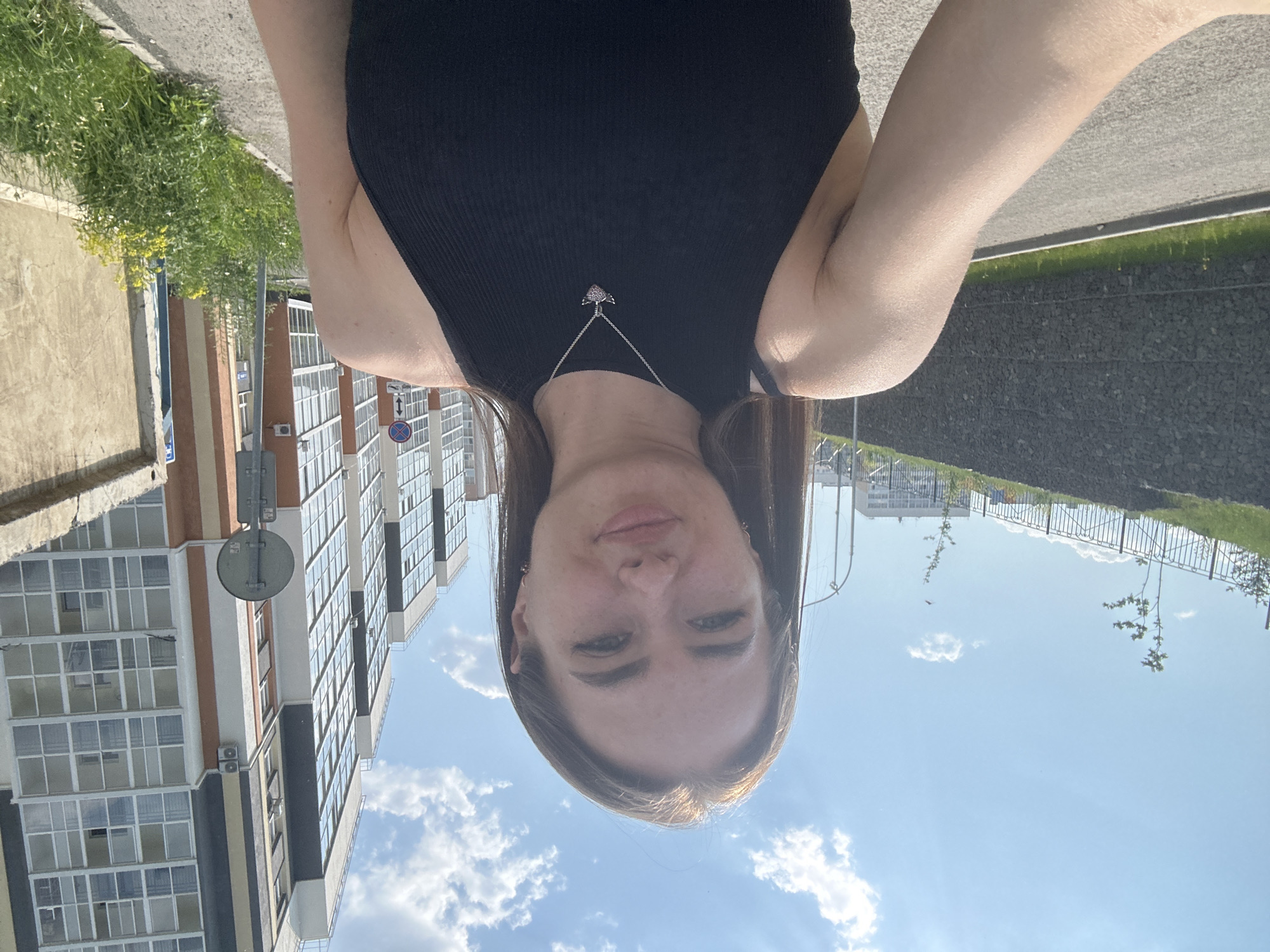 In a medium shot, a young woman with long brown hair and light skin, wearing a black tank top and a silver necklace with a heart-shaped pendant, stands outdoors. She is looking directly at the camera with a slight smile and her eyes are open. The background shows a modern cityscape with tall buildings, a blue sky with white clouds, a paved road, a stone wall, and green grass. The lighting suggests it is daytime and sunny.