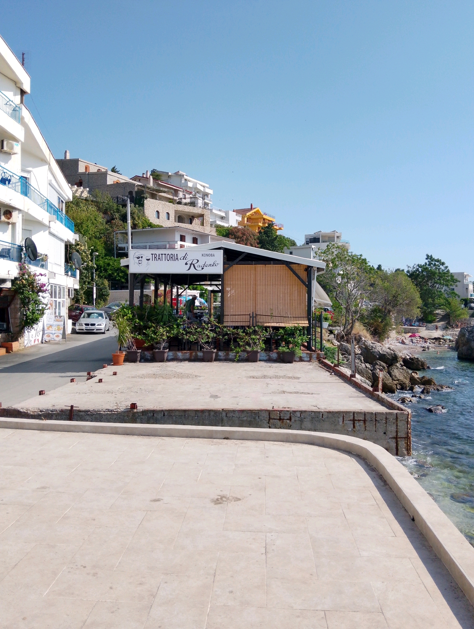 This image shows a view of the waterfront in Sutomore, Montenegro. In the foreground, there is a paved area with a curved edge bordering the sea. To the left, a white building with blue railings is visible, with a street running alongside it. A silver car is parked on the street. In the middle ground, there is an outdoor restaurant with a sign that reads "TRATTORIA di Radenko". Several potted plants are arranged around the restaurant. Beyond the restaurant, a small beach with people can be seen, along with rocky outcrops and trees. The background features a clear blue sky and houses built on a hillside. The sea is visible on the right side of the image, with clear, shallow water.