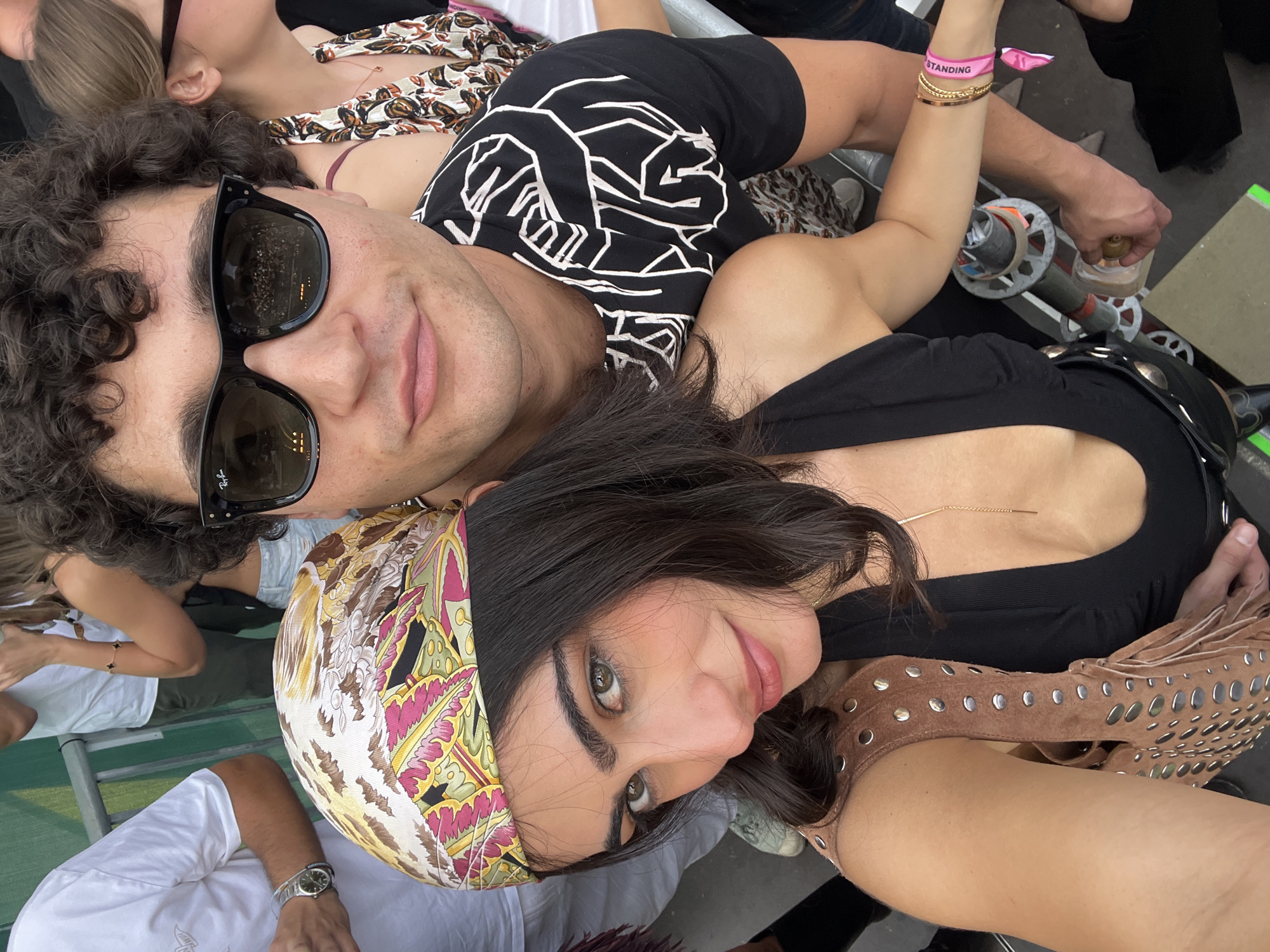 A close-up, canted angle photograph shows a man and woman posing closely. The man, with curly brown hair, wears dark sunglasses and a black t-shirt with a white abstract graphic. He has a slight smile. The woman, with dark hair partially covered by a patterned scarf, looks at the camera with a slight smile. She wears a black top and a brown, studded fringed bag or garment. The lighting is bright. Other people are visible in the background. One person is wearing a shirt with the words "STANDING". There is a metal guardrail present in the scene. The location is likely not in Madrid, Spain, as there is no indication of that place in the picture.