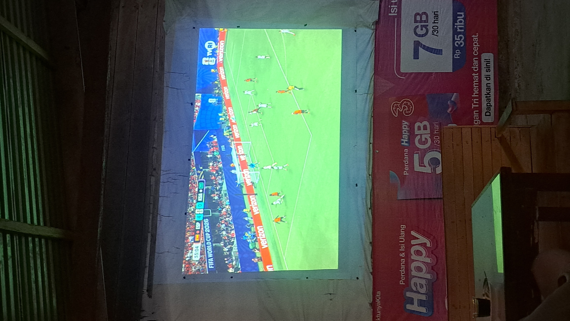 The image depicts a projector screen displaying a soccer match, likely the FIFA World Cup 2026, with the score showing ESP 4 and KSA 0 at 55:04. The match is being broadcast by TVRI. Below the screen, a red banner with white text advertises mobile data plans from "Happy" and "3" providers, offering 5GB and 7GB of data for 30 days, with prices listed in Indonesian Rupiah. The scene appears to be in an informal setting, possibly an outdoor viewing area or a cafe, as indicated by the presence of a wooden table with a glass of dark liquid and some seating. The overall environment suggests a casual gathering to watch the game.
