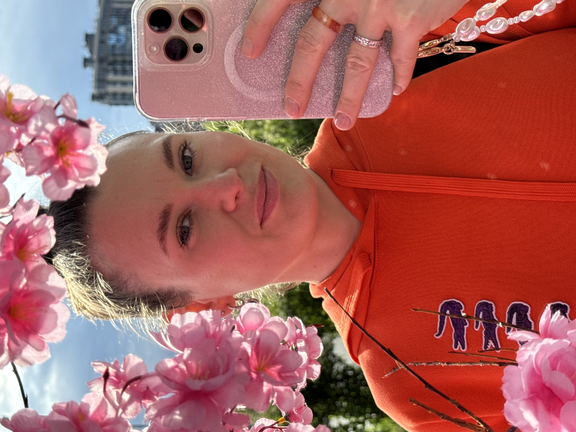 A young woman with blonde hair pulled into a bun takes a selfie in front of a building. She is wearing an orange hooded sweatshirt with a purple alligator logo. She is holding a pink smartphone in her right hand and is wearing two rings on her right index finger. The background shows a blue sky with clouds and a building, possibly in Saint Petersburg, Russia. Pink flowers frame the left side of the image.