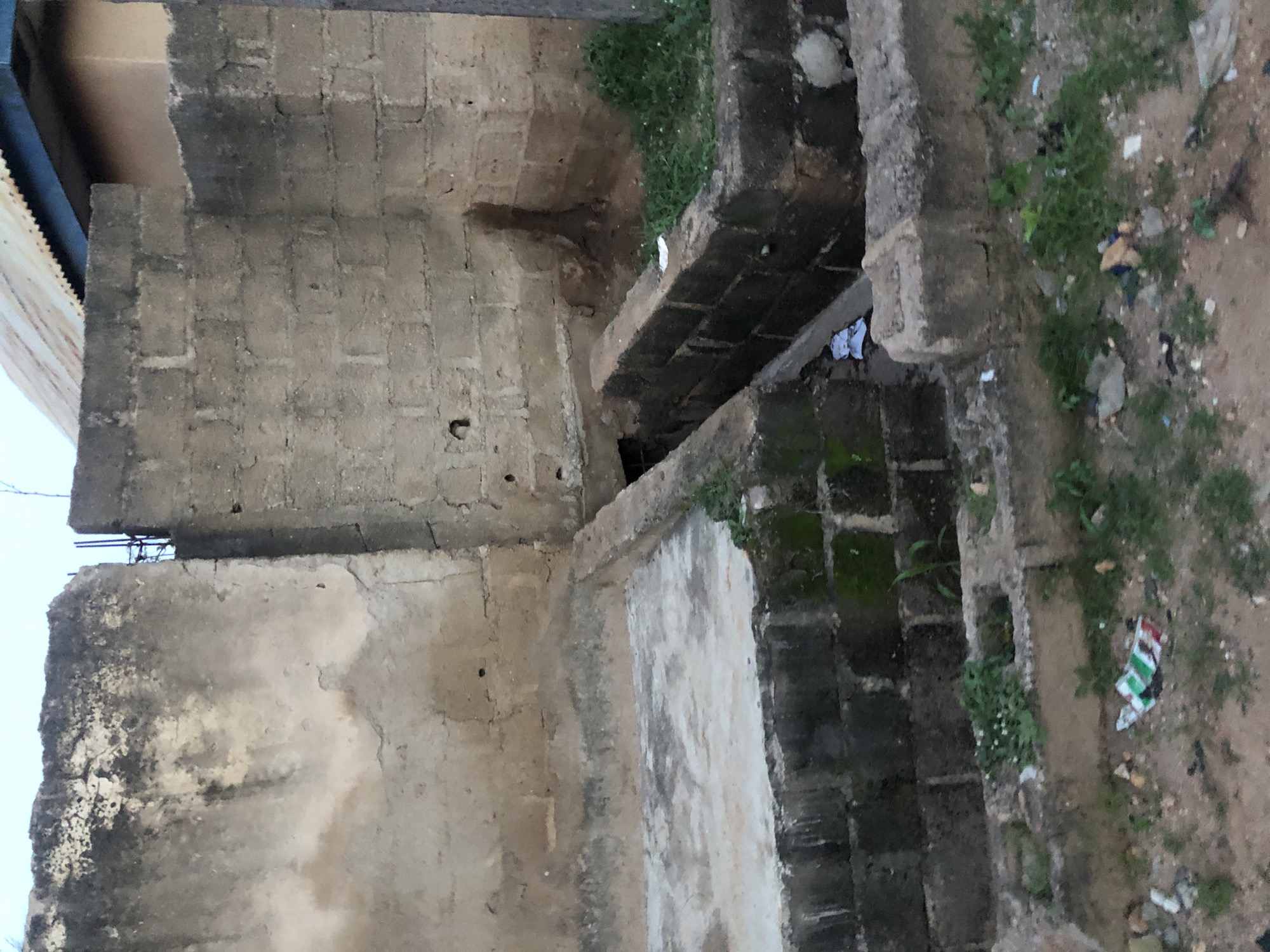 The image depicts an outdoor scene in Jalingo, Nigeria, focusing on a dilapidated drainage system and surrounding structures. The foreground is a rough, uneven ground covered in dirt and scattered debris, including colorful wrappers, a small plastic bag, and other unidentifiable objects. Several small green plants are growing sporadically from the ground and from within the drainage structure.
The central focus is a brick-lined drainage channel, partially filled with water and debris. The bricks are dark and appear weathered, some showing signs of moss growth. The channel leads into a dark opening in a larger concrete structure.
Flanking the drainage are low, rough-hewn concrete walls and a raised, roughly plastered concrete platform. To the left, a larger, weathered concrete wall dominates the frame. This wall is textured and stained, showing patches of discoloration and erosion. On the right, a section of a building with a corrugated metal roof and a lighter-colored wall is visible, suggesting proximity to residential or commercial structures. The overall impression is one of neglect and disrepair, characteristic of informal settlements or areas undergoing development. The sky above is overcast, with a pale, diffused light.