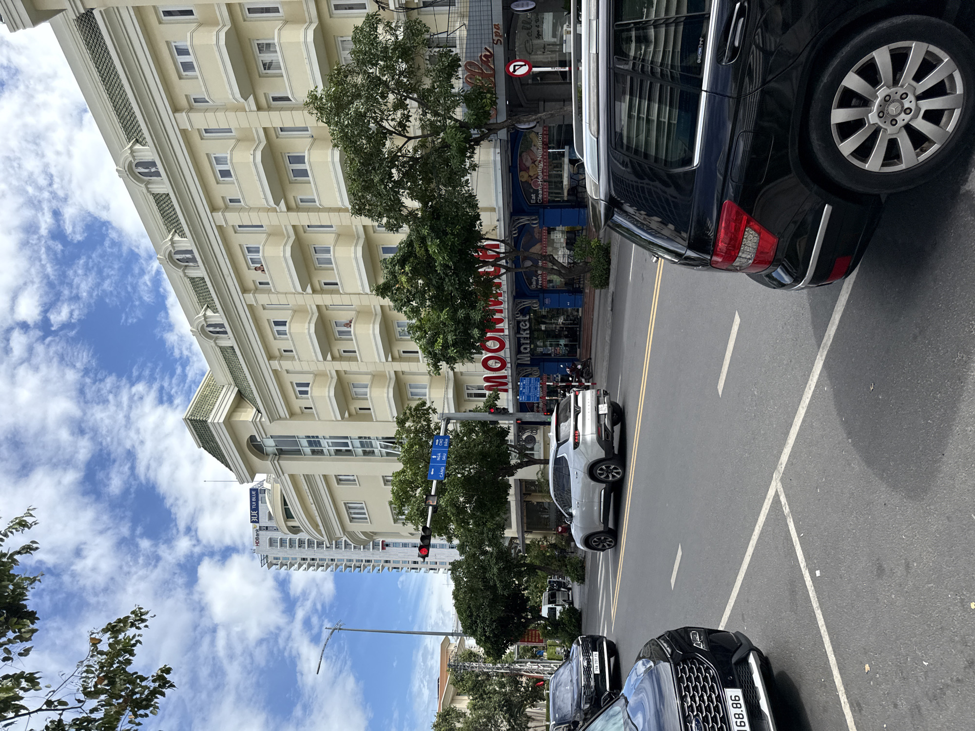 A street scene in Nha Trang, Vietnam, captures a moment with several vehicles, buildings, and a traffic signal under a partly cloudy sky. In the foreground, the rear of a black Mercedes-Benz SUV is visible on the right side of the frame, facing away from the viewer. To its left, a silver SUV is moving forward in the street, and a black SUV with a license plate reading "79A-257.86" is stopped at an intersection. A white van is also seen further down the street.
Dominating the background is a multi-story, cream-colored building with arched windows and green-tiled roofs. A large sign for "MOON MARKET" is prominently displayed on the building's facade, with additional signage for "Galla Spa." To the left of this building, a tall, slender skyscraper is visible. The street is paved with asphalt, marked with yellow and white lane lines. A traffic light hanging over the intersection shows a red signal. Trees line the street, and various signs are posted, including directional signs indicating "CANG SAU DAM" and a "no right turn" symbol. The overall environment suggests a commercial and residential area within the city.