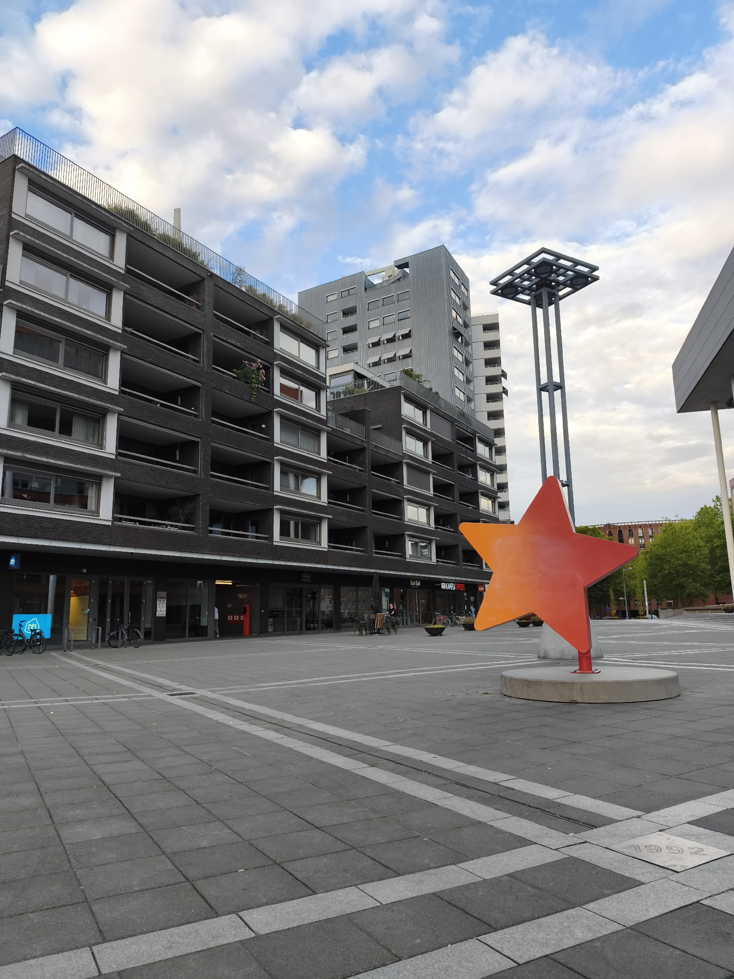 The image shows the city of Maastricht, Netherlands, specifically the Sint Maartenslaan area, captured on a cloudy day. In the foreground, a large, orange-red star sculpture stands on a concrete base. Behind the sculpture, a modern, multi-story dark brick building with balconies and numerous windows dominates the left side of the frame. Further back, a taller, grey, modern building is visible, and to the right of the star, a large, dark, angular structure juts into the frame, likely part of another building. The ground is paved with grey square tiles and lined with light grey rectangular strips. A metal pole with a square, grid-like top rises behind the star sculpture. The sky is partially filled with white, scattered clouds. Bicycles are parked near the entrance of the dark brick building. The overall scene depicts an urban public space with contemporary architecture.