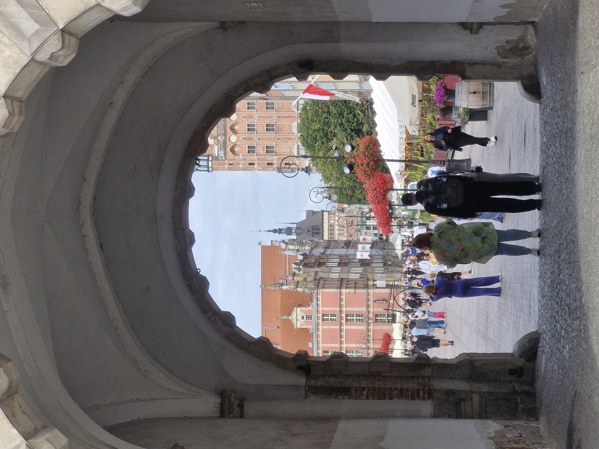 An eye-level, medium shot captures a street scene in Gdańsk, Poland, viewed from beneath a large, weathered stone archway. The archway, constructed of roughly hewn stones and featuring a decorative carved edge, frames a bustling city square beyond.
In the foreground, partially obscuring the view, stand two figures facing away from the camera. The person on the left is a man wearing a black backpack and dark clothing. To his right stands a woman wearing a light green, patterned jacket over blue pants. They are positioned on a cobblestone surface that transitions to a paved walkway.
Beyond the archway, the square is filled with people walking in various directions. The architecture is a prominent feature, with colorful, ornate buildings lining the street. A prominent red-roofed building with a stepped gable and statues is visible. Further down the street, a tall brick tower with arched windows stands against a pale sky. Red flowers in hanging baskets and planters add vibrant splashes of color to the scene, and white awnings of outdoor cafes are visible. A Polish flag, red and white, hangs from a pole. The overall impression is of a lively public space on a bright, possibly overcast day.