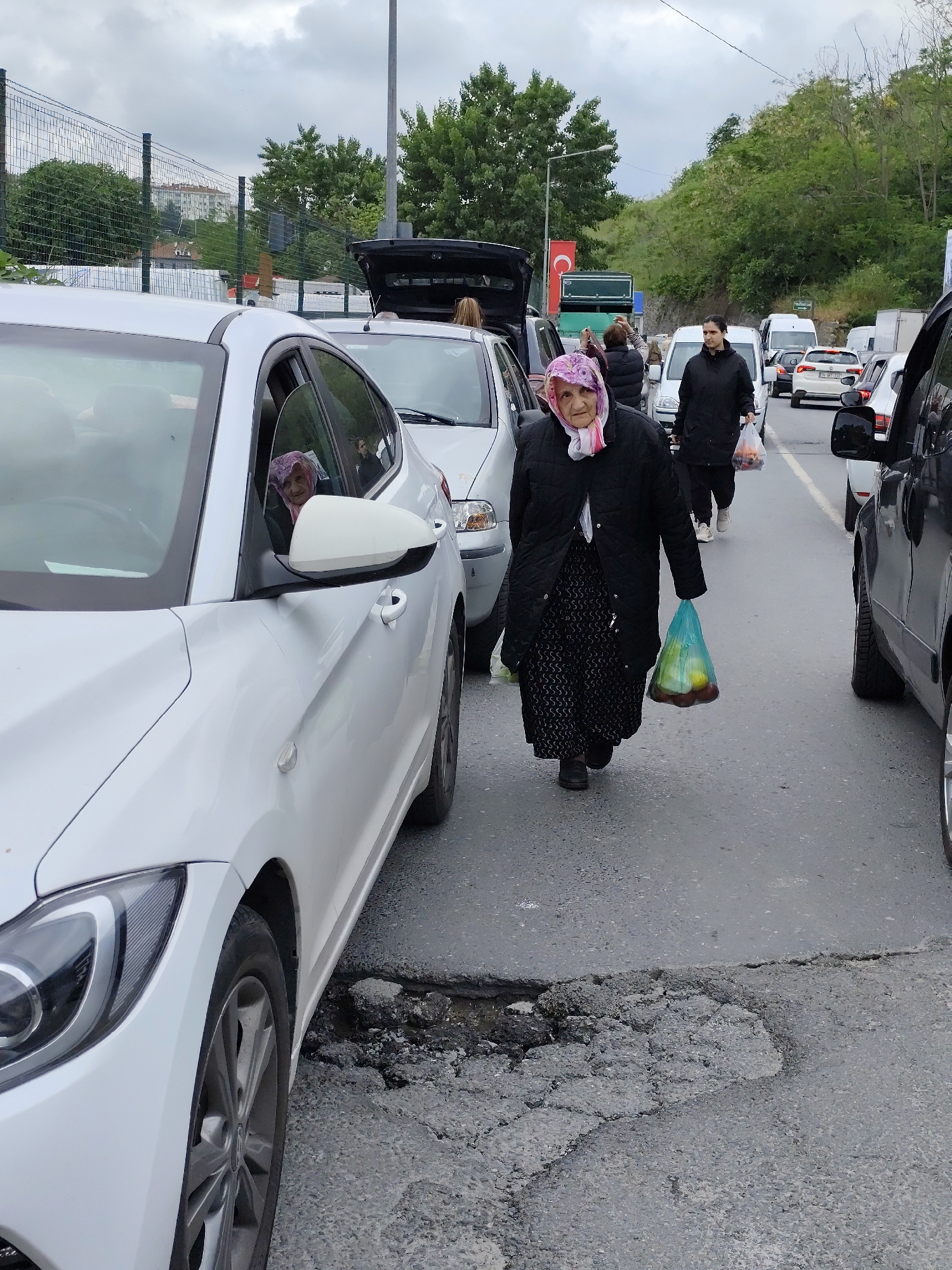 A medium-angle, eye-level shot depicts a woman in traditional garb walking in the middle of a road amidst slow-moving car traffic.
The main subject is a woman, likely elderly, wearing a dark floral dress covered by a long black coat, and a pink and white patterned headscarf. She holds a translucent blue plastic bag filled with what appears to be fruits. She is walking towards the viewer.
To the left of the woman is a white sedan. A woman with a similar headscarf is visible in the driver's seat. To the right is a black SUV. Behind the main subject, the road is filled with more cars, some with their trunks open. There are other pedestrians visible, one in a black coat carrying a bag.
The environment appears to be an urban area. There are trees and buildings visible in the background, along with a Turkish flag. The road itself is grey and weathered, with a noticeable pothole near the white car. The sky is overcast.