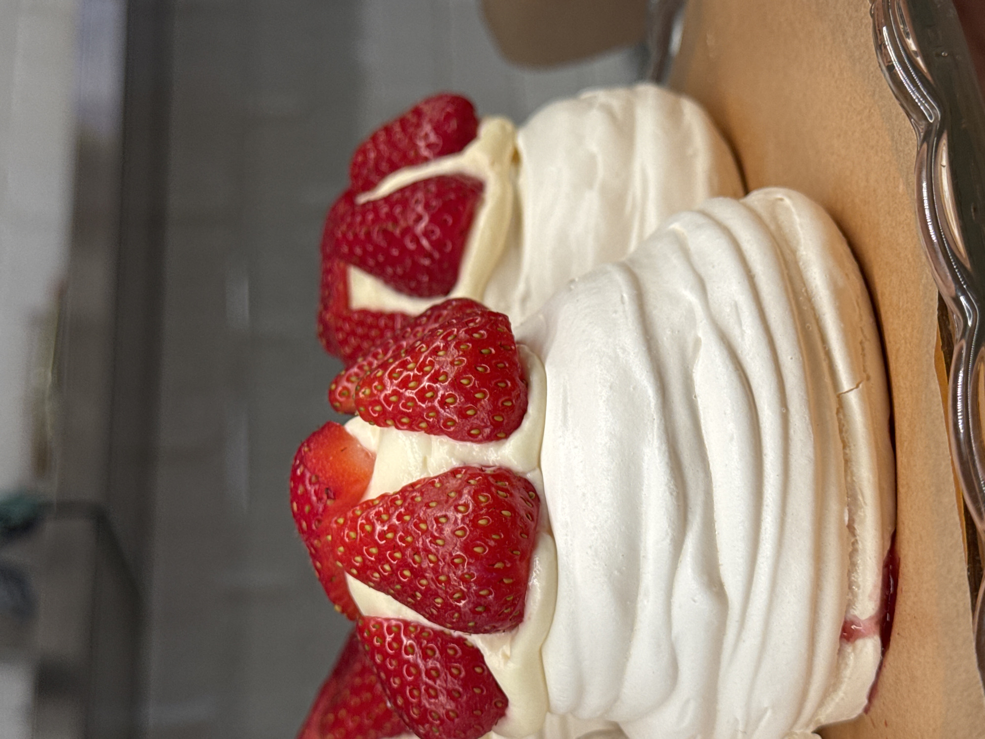 This close-up image, presented on its side, features a dessert composed of white meringue or pavlova bases, fresh red strawberries, and creamy filling. Multiple individual dessert portions are visible, each with a distinct white meringue shell exhibiting a textured, ridged surface and slight cracking. Nestled between these meringue components and serving as a bed for the fruit is a generous amount of creamy, off-white or light yellow filling, possibly whipped cream or custard.
Bright red, ripe strawberries, some appearing whole and others possibly halved, are arranged abundantly on top of the cream, showcasing their textured skin and seed patterns. A silver-colored, scalloped metal serving tray is partially visible on the right, supporting the dessert, which rests on light brown parchment or baking paper. Near the bottom right, a faint reddish liquid, likely strawberry juice, has seeped onto the parchment.
The background is uniformly blurred and dark gray, suggesting an indoor setting such as a kitchen or bakery, but without specific identifying features. The image focuses solely on the dessert's presentation. No specific activity or event beyond the display of the food is depicted. There are no visible location cues within the image that point to Moscow, Russia, nor are there any indications of the time of day, weather, or explicit emotions. No text is visible in the media file.
