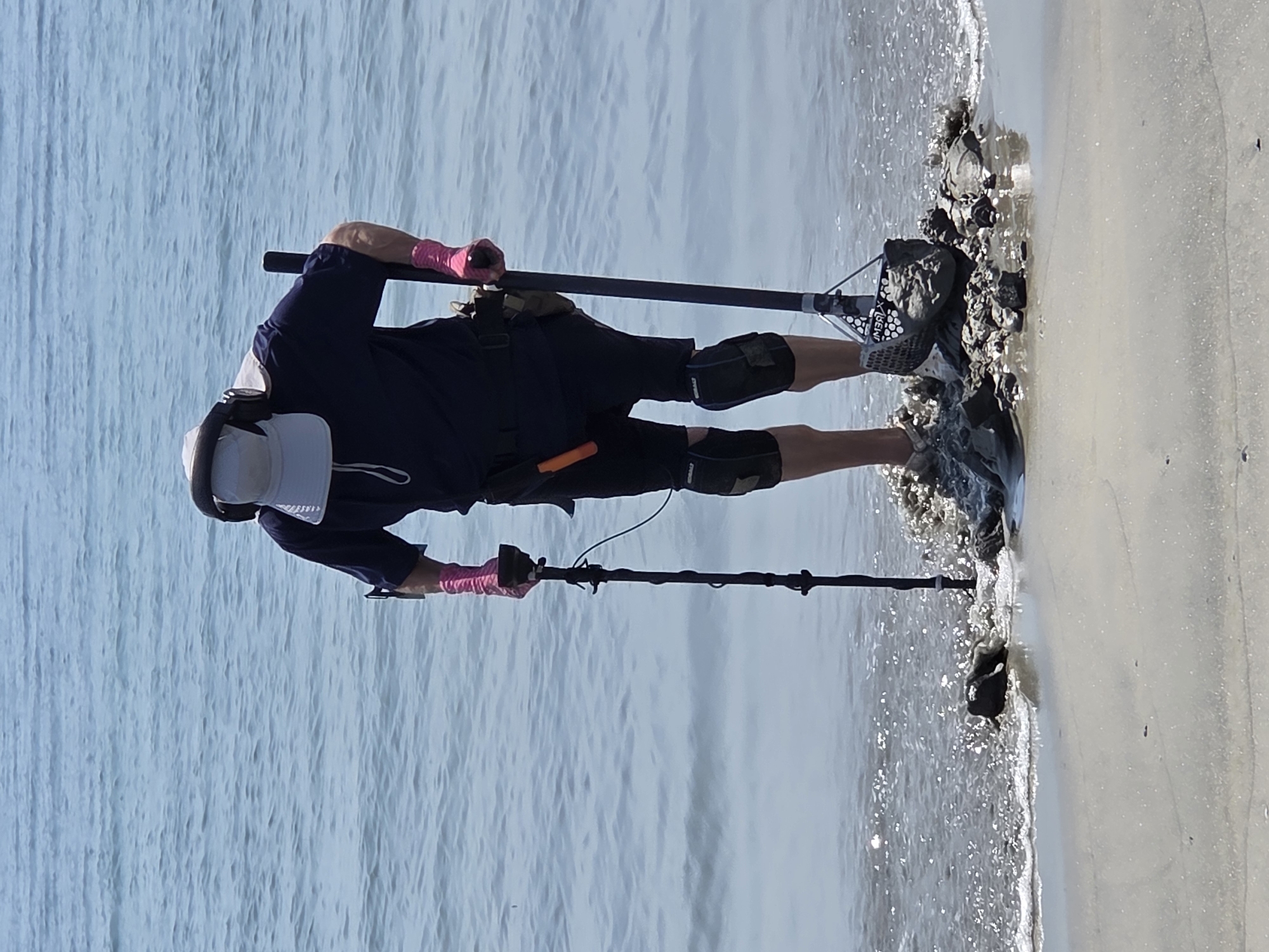 A person stands in the shallow water at the edge of the ocean, at a beach in Long Beach, United States of America, with a metal detector. The person is wearing a dark blue t-shirt, dark shorts, knee pads, a white wide-brimmed hat with a grey band, headphones, and bright pink gloves. They are holding a metal detector in their right hand, which has a search coil at the bottom. A shovel with a mesh basket is attached to the metal detector pole, and it contains some wet sand and rocks. There is a tool with an orange handle attached to a belt around the person's waist. The water is calm and blue, with some small waves breaking on the shore. The sand is wet and appears to be mixed with small rocks and shells where the person is standing. The lighting suggests it is a sunny day.