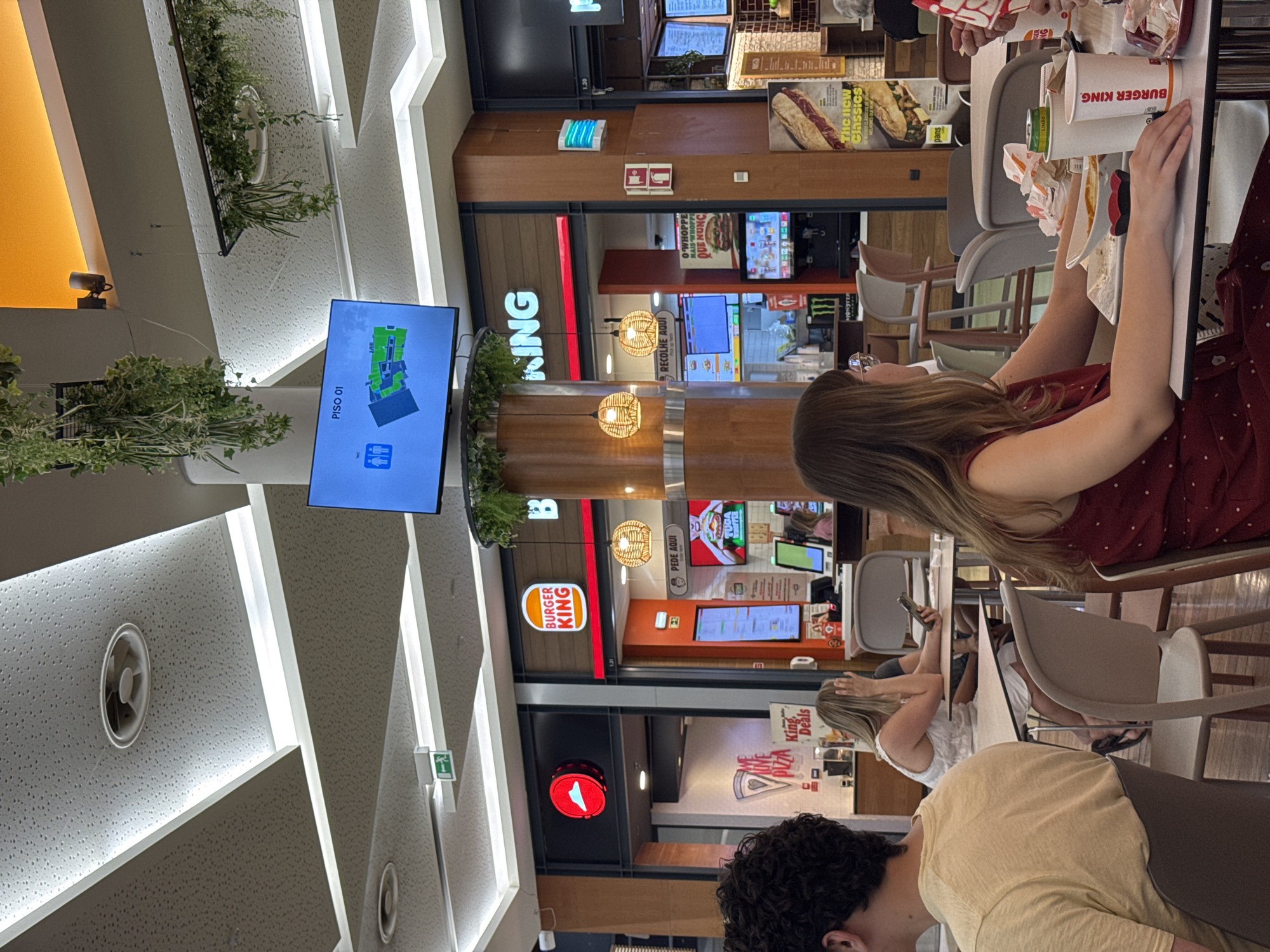 The image depicts the interior of a Burger King restaurant in Castelo Branco, Portugal, specifically within a shopping center, as indicated by the presence of multiple food establishments and the mall-like ceiling and lighting. The perspective is from a dining area, looking towards the counter and kitchen area of the Burger King. In the foreground, a woman with long brown hair wearing a red dress is seated at a table, her hands resting on the table. To her left, another person's arm is visible, holding a white cup with the Burger King logo. Partially visible to the left foreground is a person with curly dark hair wearing a yellow t-shirt. Behind the woman in the red dress, a blonde woman in a white dress is seated at another table, holding a smartphone and looking down.
The Burger King storefront is prominently displayed with its signature orange and red branding. Inside the restaurant, order screens are visible, along with a menu board displaying food items. A Pizza Hut storefront is adjacent to the Burger King. The environment is well-lit with artificial lighting from recessed ceiling fixtures and decorative pendant lamps. Green hanging plants are visible, adding a touch of greenery to the indoor setting. A digital screen displaying a floor plan with icons for elevators and restrooms is mounted on the wall above the counter area. The overall scene suggests a busy food court environment within a commercial complex.