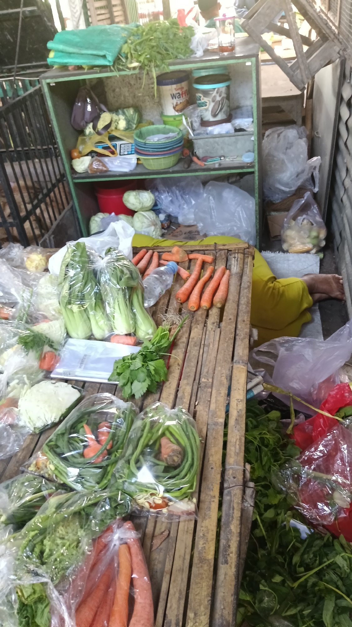 This is a medium-shot, eye-level, indoor photograph of a stall at a market in Purwokerto, Indonesia. The stall is cluttered with fresh produce, some of which is packaged in plastic bags. In the foreground, there are several bunches of green leafy vegetables, including what appears to be bok choy and possibly kale. There are also carrots, some whole and some sliced, displayed on a bamboo mat. To the left of the stall, a person wearing a yellow garment is lying down, their legs visible, suggesting they may be resting or sleeping. Behind the vegetables, a green metal shelf unit holds various items, including containers of what appear to be snacks, bowls, and what looks like a small scale. A red bucket is also visible on the lower shelf. The overall impression is of a busy, somewhat disorganized market stall.