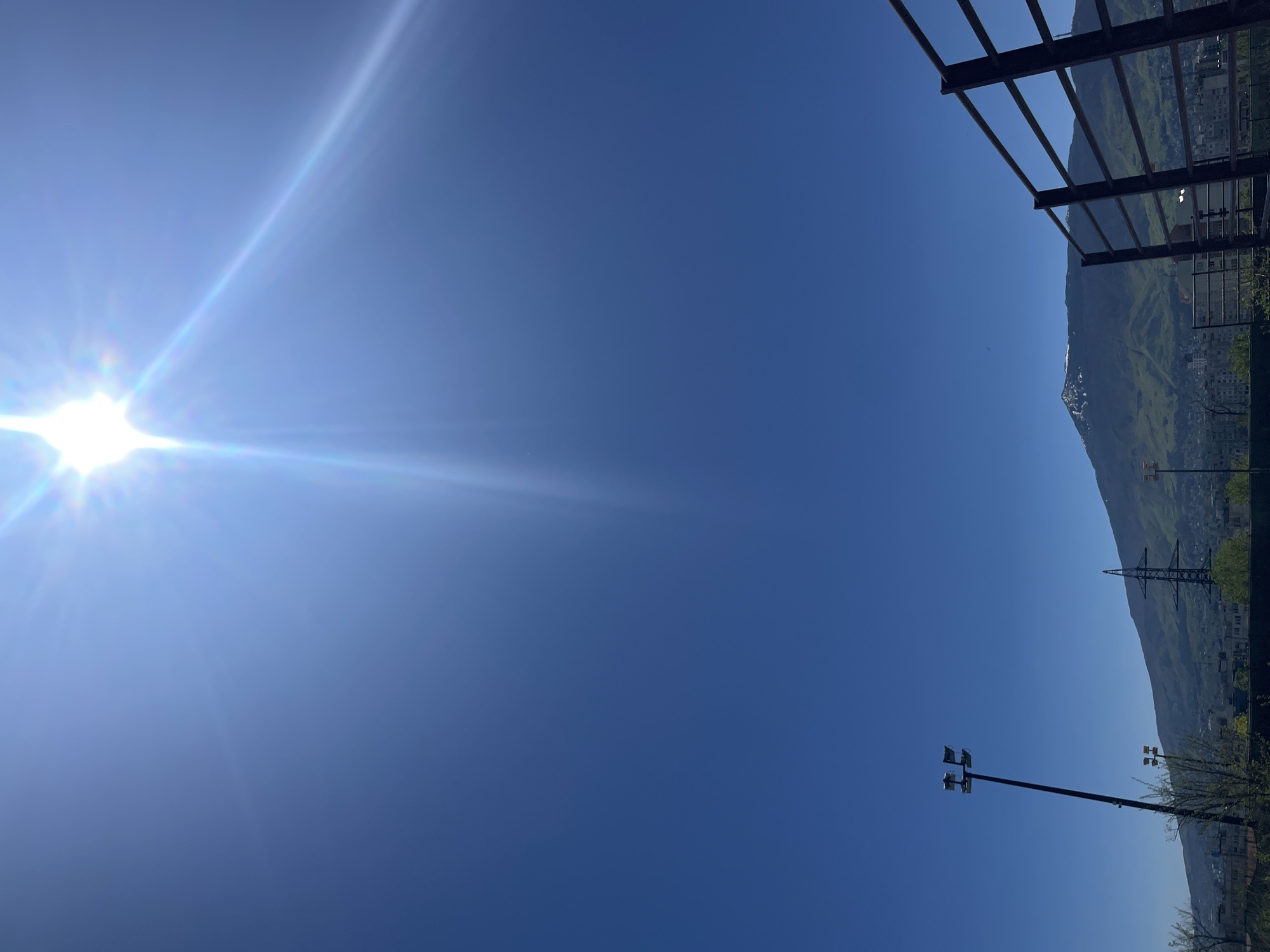 The image shows a clear, blue sky with the sun shining brightly in the upper left corner. To the right of the sky, there is a mountain range visible, with some snow on the peaks. In the lower right corner, there are trees, light poles, buildings and power lines. The location is inferred to be in Sofia, Bulgaria. The frame suggests an urban environment bordering on natural terrain. In the upper right corner there is an ironwork construction.