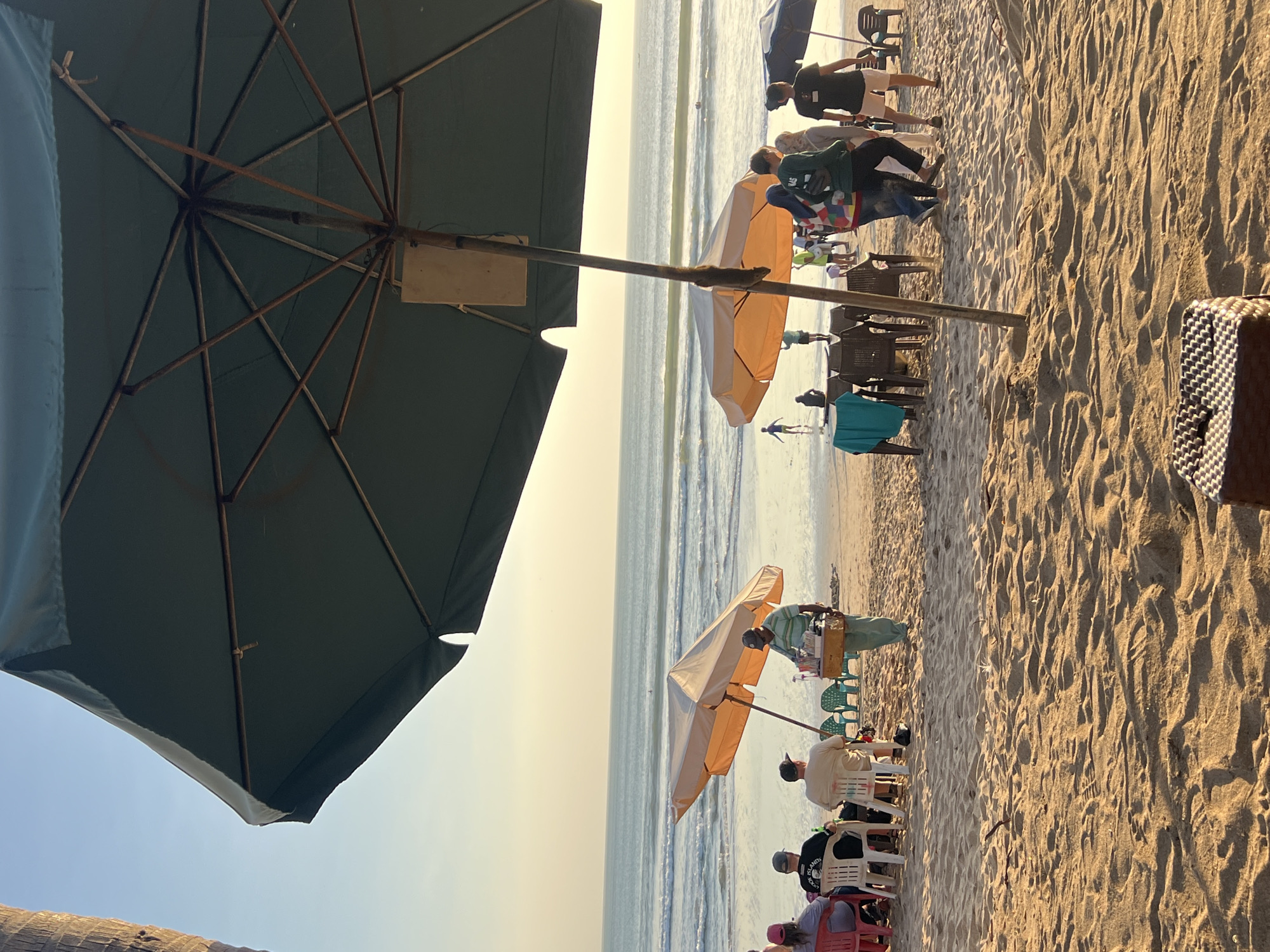 In Kuta, Indonesia, a beach scene unfolds under a hazy sky. A large, dark green umbrella dominates the foreground, casting a shadow over a sandy beach. Two smaller, beige umbrellas are set up further down the shore, providing shade for several people seated on chairs. A vendor, wearing a striped shirt and a cap, stands near one of the beige umbrellas, carrying a tray of drinks. In the background, the ocean stretches to the horizon, with gentle waves lapping the shore. Several other people are scattered across the beach, some standing, others walking. The overall atmosphere is one of a relaxed day at the beach, with a touch of commercial activity.