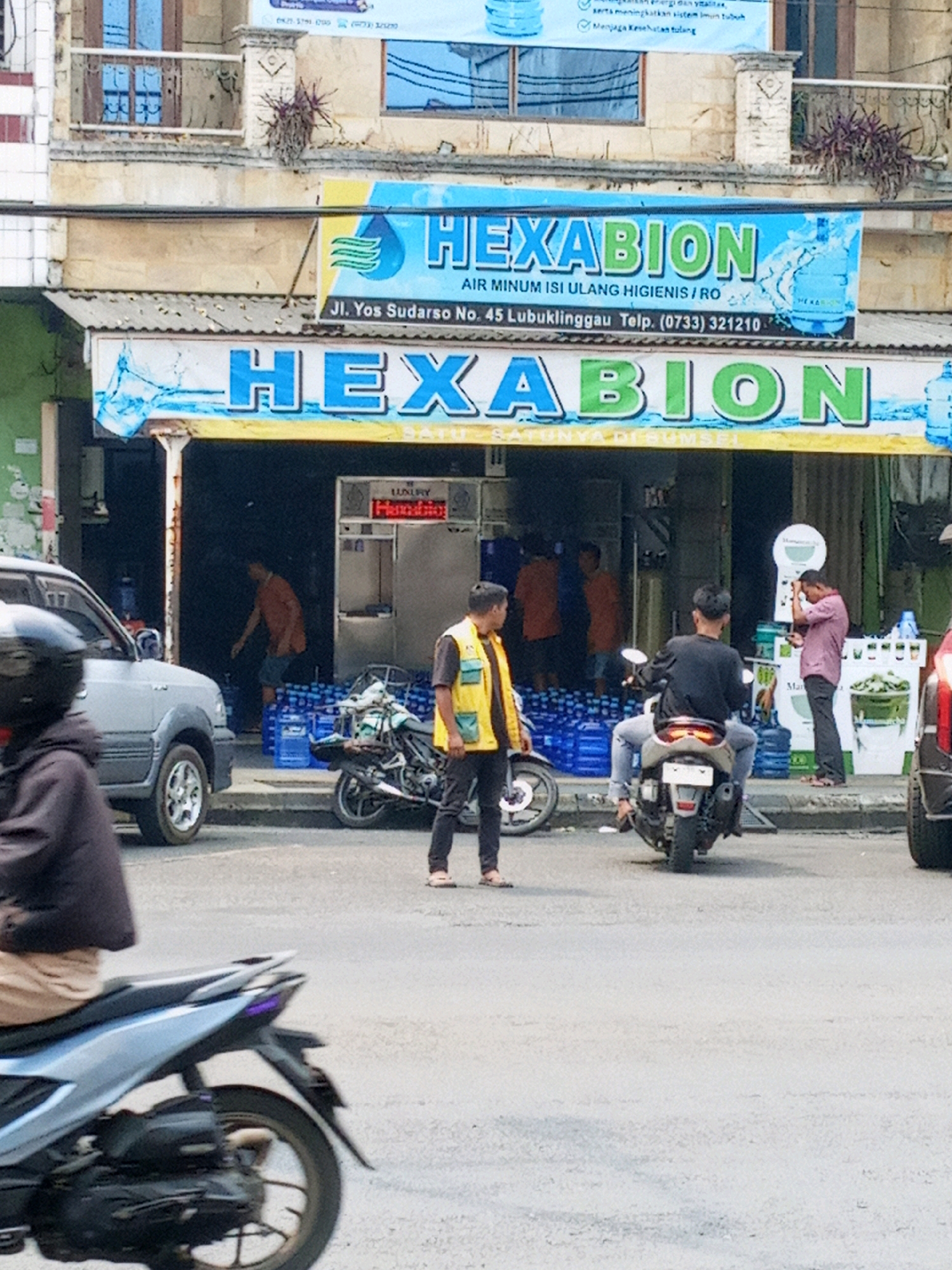 The image captures a street scene in Lubuklinggau, Indonesia, featuring a storefront for "HEXABION," an establishment selling water. The storefront is adorned with two large banners displaying the HEXABION logo and advertising "AIR MINUM ISI ULANG HIGIENIS / RO" (Hygienic Refill Drinking Water / RO). The address "Jl. Yos Sudarso No. 45 Lubuklinggau Telp. (0733) 321210" is visible on the upper banner.
In front of the store, a man in a yellow vest and dark pants is standing beside a motorcycle. Another man, wearing a black shirt and grey pants, is seated on a scooter. Several large blue water jugs are stacked outside the store, indicating their retail nature. A silver SUV is parked to the left of the frame, and another vehicle, partially visible on the far right, is also present. Inside the store, through the open entrance, more water jugs are visible, and a person in an orange shirt can be seen. The overall environment suggests a commercial area on a street in Lubuklinggau.