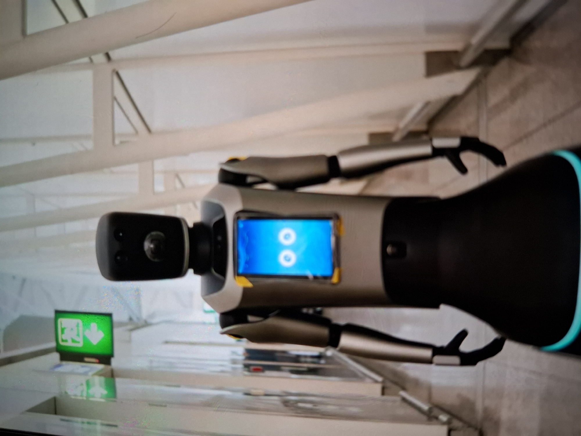 In an indoor environment, possibly an airport terminal or a similar public space with white structural beams and light-colored walls, a humanoid robot stands facing forward. The robot has a black, rectangular head with two small circular indentations above a camera lens. Its body is primarily silver with black accents. A rectangular screen on its torso displays two large, white circular "eyes" on a blue background. The robot has articulated arms and hands. To the left of the robot, a green exit sign with a white arrow pointing left is visible on the wall. The floor appears to be a polished surface, reflecting some of the ambient light.