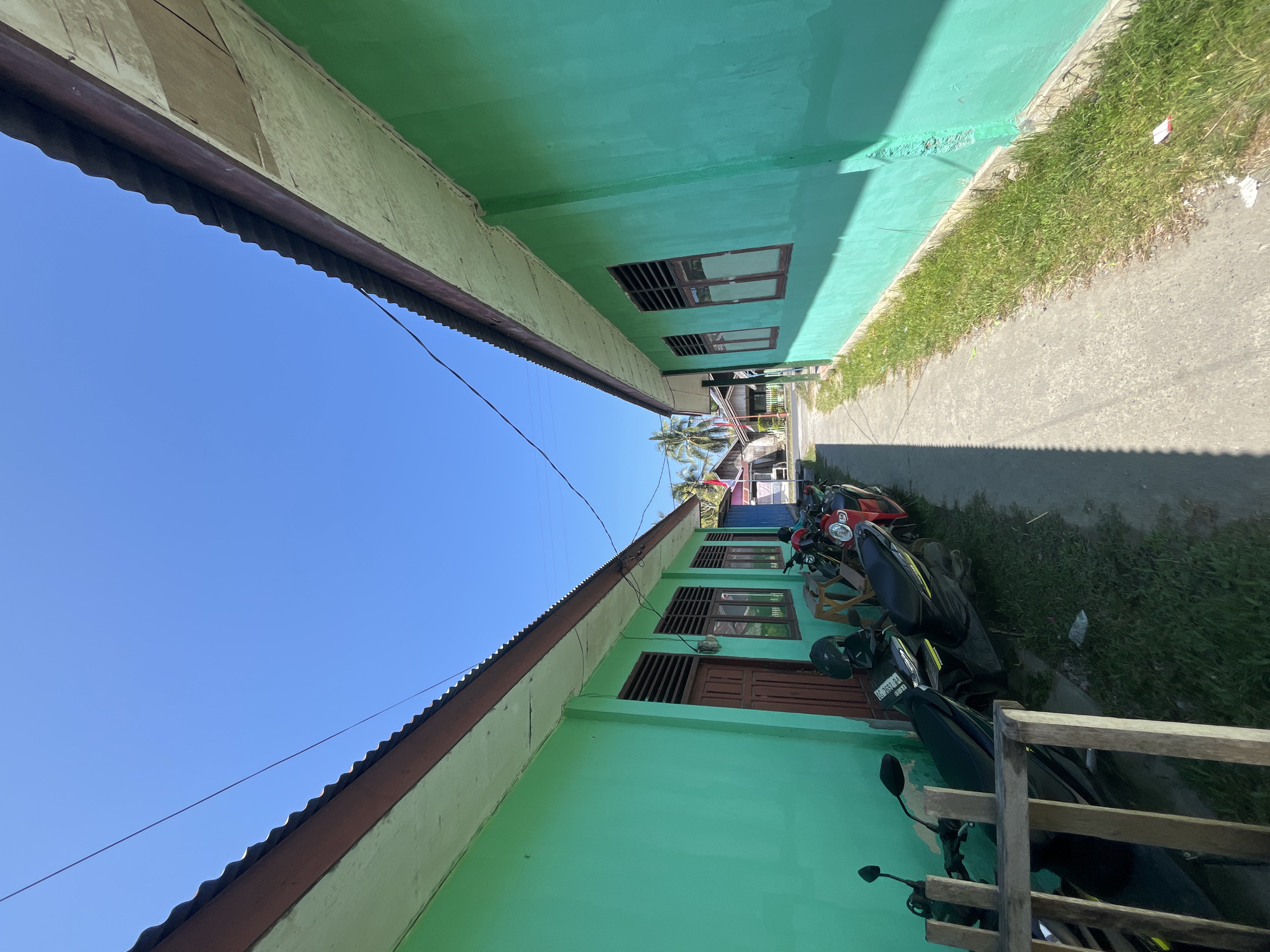 This is an eye-level, wide-angle, outdoor shot of a narrow walkway between two teal-colored buildings in Sanana, Indonesia. The sky is clear blue, and the sun is casting shadows. On the left side of the walkway, several motorcycles are parked along the building, some with helmets on them. The building on the left has a brown wooden door and several windows with brown frames and louvers. The building on the right also has windows with brown frames and louvers. The walkway itself is made of concrete and is flanked by a strip of green grass on the right. In the distance, other buildings are visible, along with palm trees.